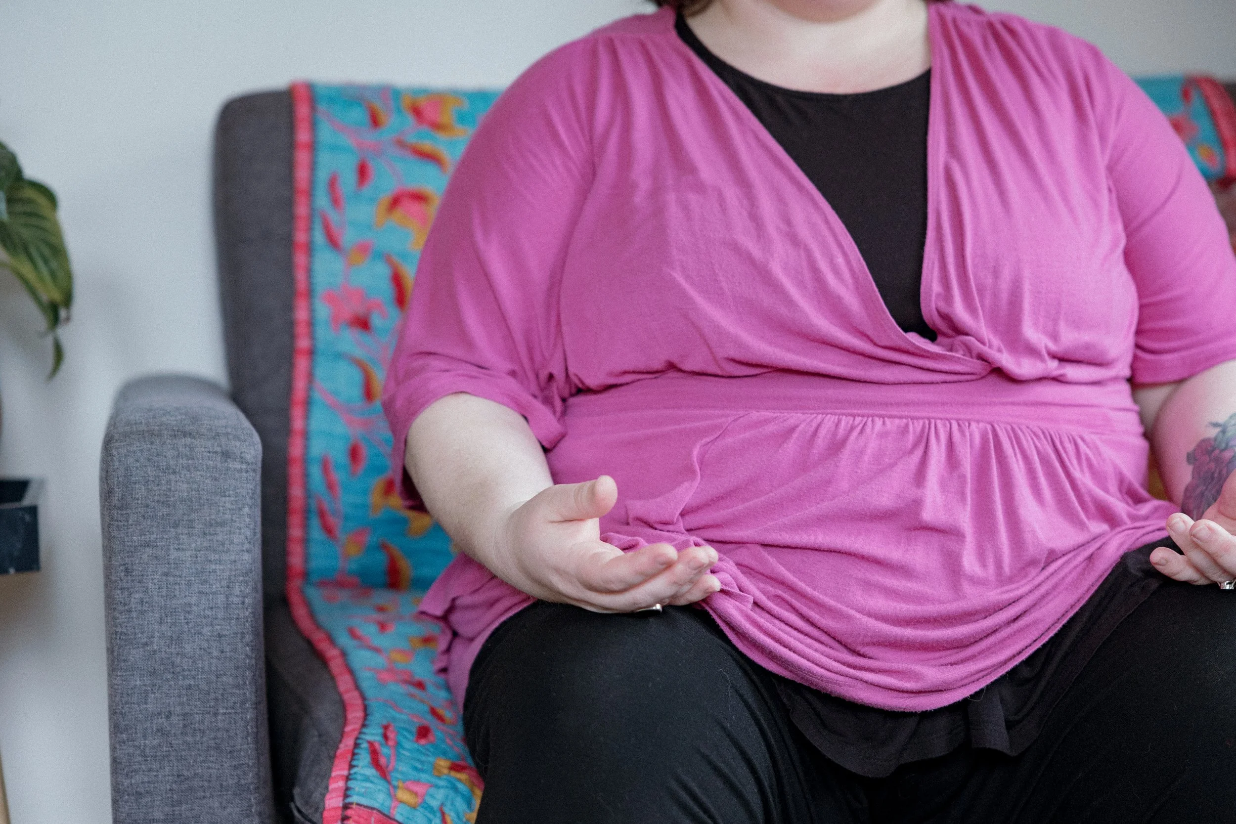 A woman sitting on a gray couch with colorful pillow, wearing a pink top and black underlayer, with her hands resting on her lap.