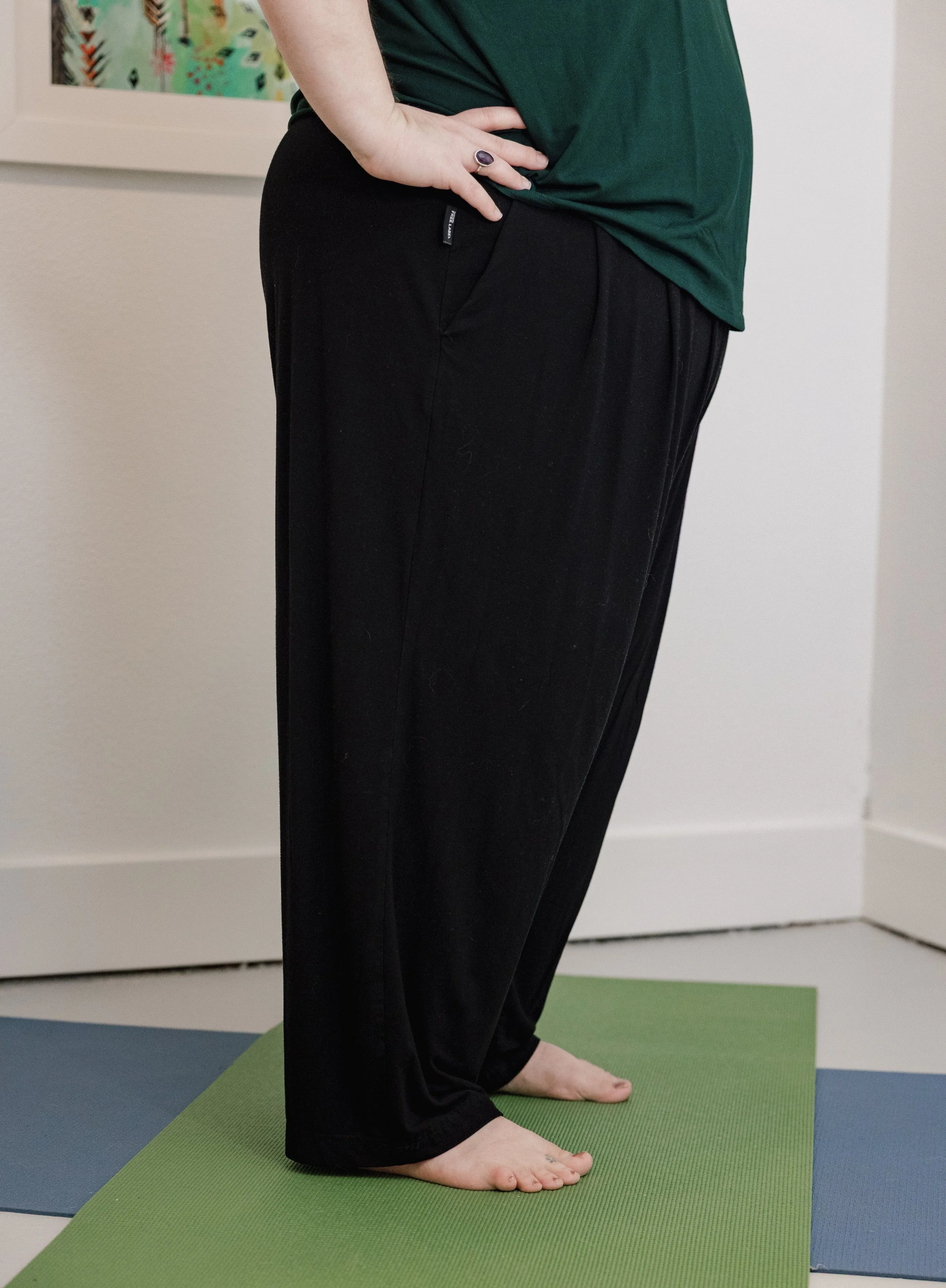 A person standing on a green yoga mat, barefoot, wearing black pants and a dark green top. Their hand, with a ring on the index finger, is resting on their hip, and they are in a yoga pose indoors with colorful artwork on the wall behind them.