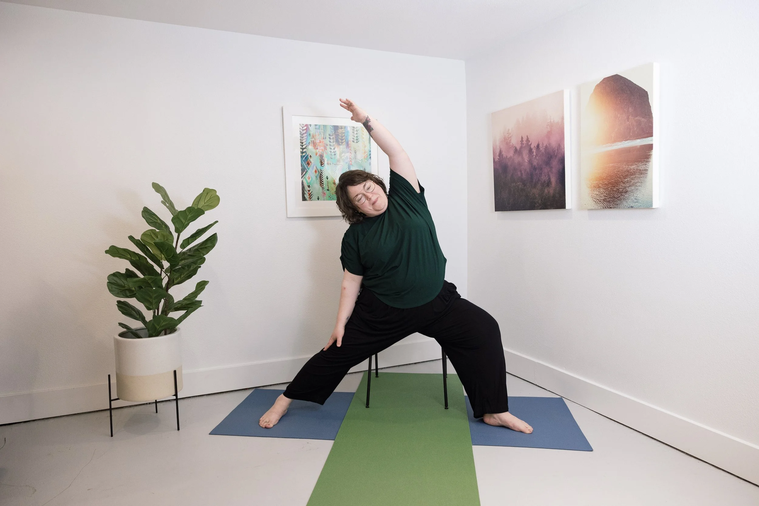 Person practicing yoga indoors on blue and green mats, standing in a wide-legged pose with one arm extended overhead and the other touching the leg, near a potted plant and colorful artwork on the wall.