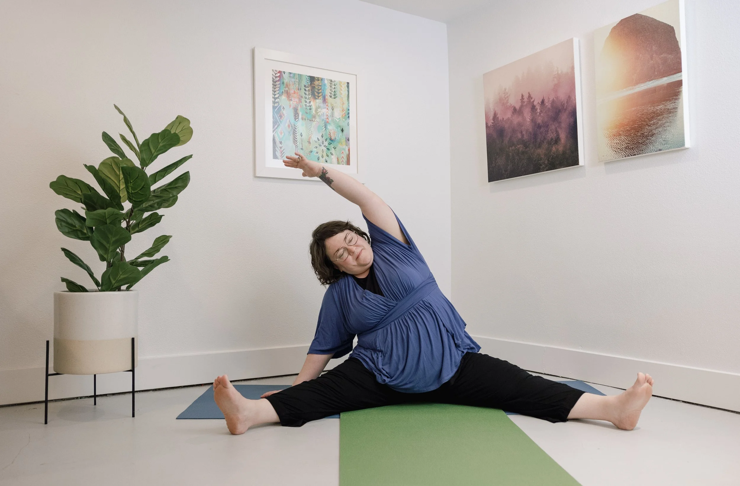 A person practicing yoga indoors, performing a side stretch pose on a green yoga mat. They are wearing glasses and a blue top, with a large green potted plant nearby. Three landscape paintings hang on the white wall behind them.