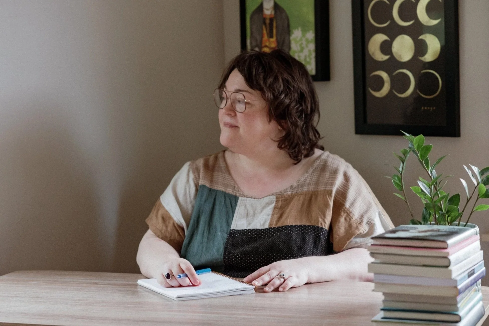 A woman with short, wavy brown hair and glasses sitting at a desk with stacked books, writing in a notebook with a blue pen. There is a green potted plant on the desk and framed artwork on the wall behind her.