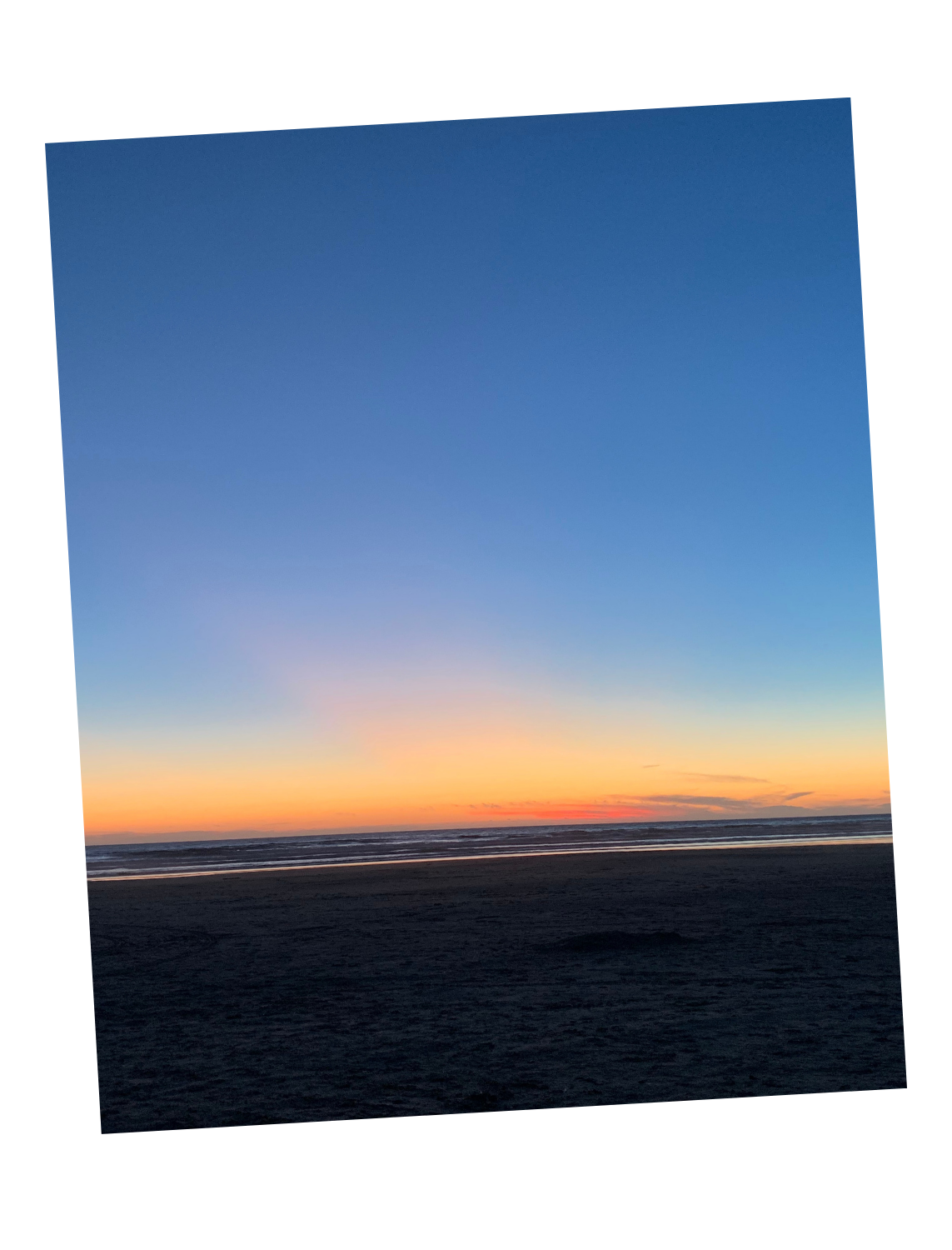 Sunset over a beach with dark sand, gentle waves, and a colorful sky with hues of orange, yellow, pink, and blue.