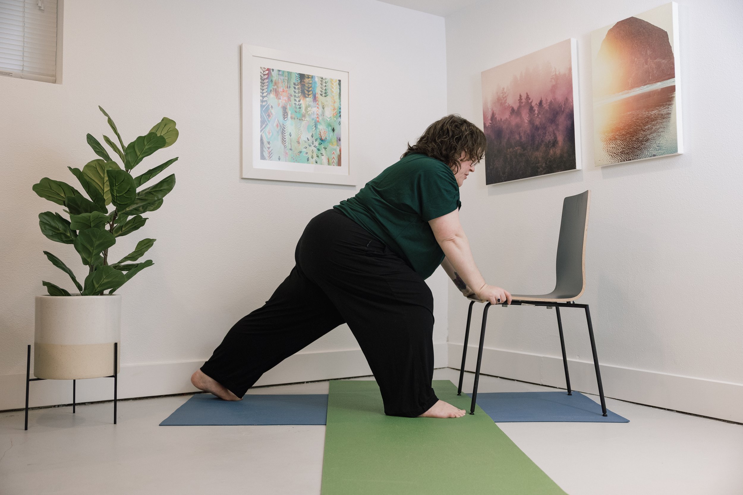 A woman in a green shirt and black pants doing a yoga pose on a green and blue mat in a room with white walls. The room has a potted plant in a white planter, and three framed nature-themed artworks hanging on the wall. She is holding a chair for support while performing a stretching or yoga pose.