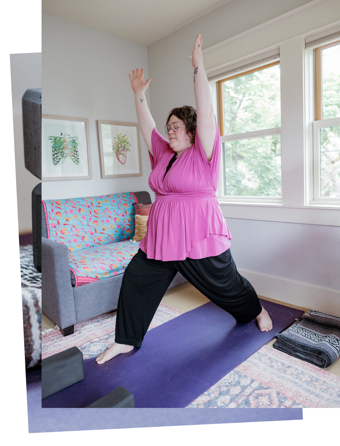 A person practicing yoga in a room with large windows, sitting on a purple yoga mat in a Warrior pose with arms raised.
