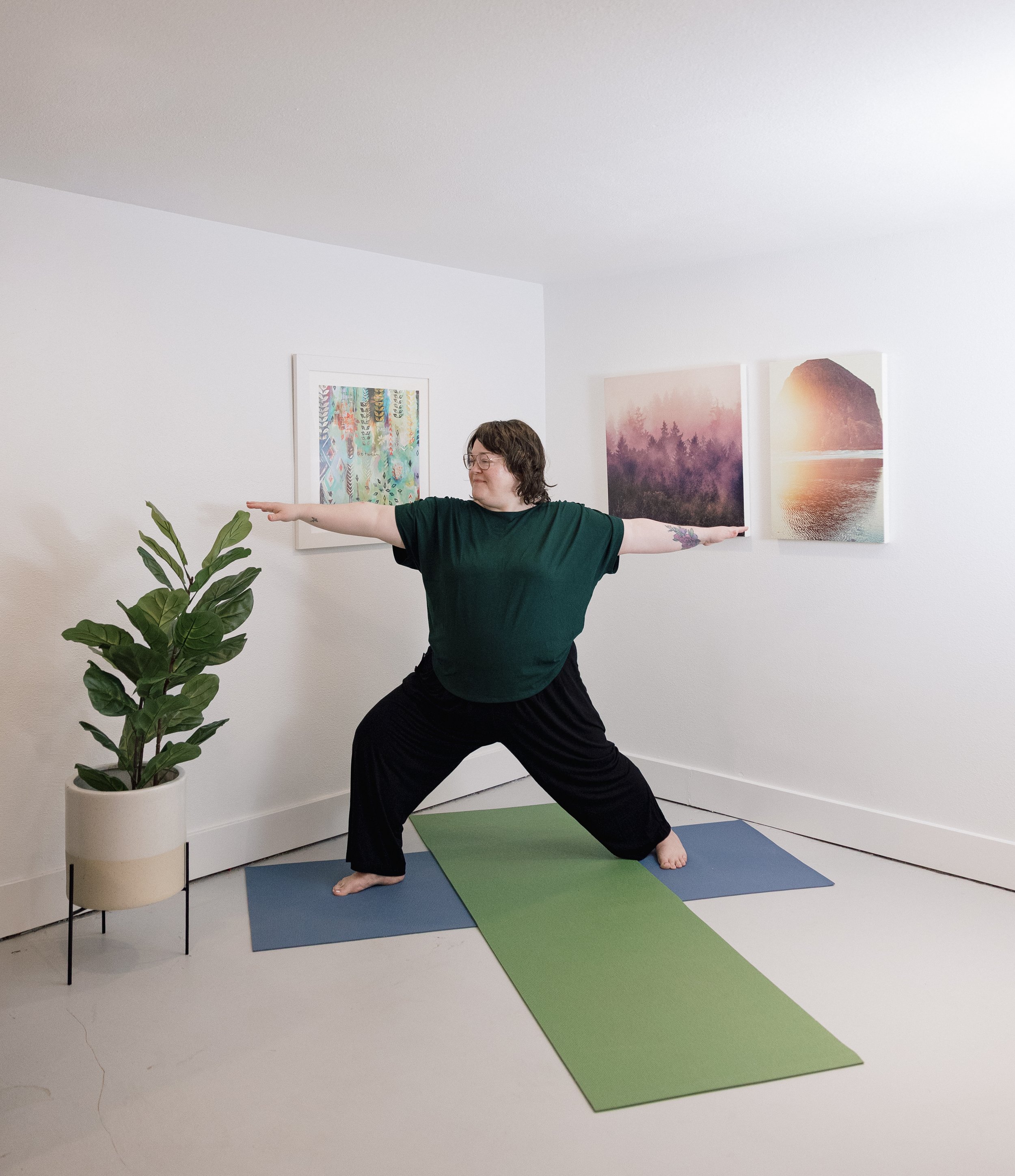Woman practicing yoga in a living room with minimal decor. She is standing on a multicolored yoga mat, extending her arms, with artwork on the wall and a large potted plant nearby.