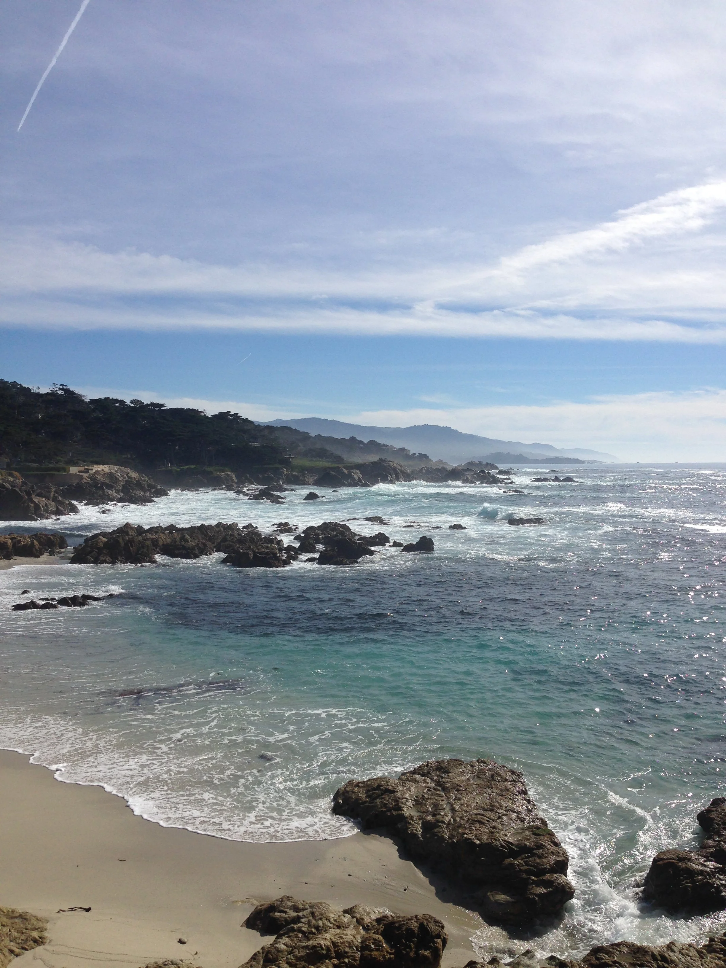 A scenic beach with white sand, rocks in the foreground, and waves crashing on the shore under a partly cloudy sky.