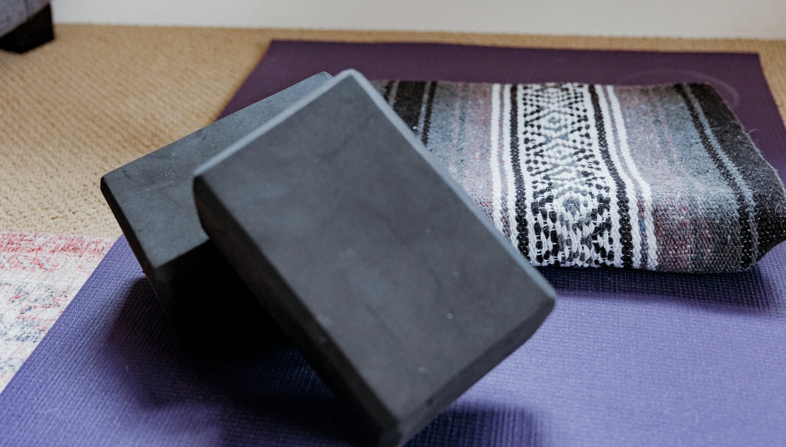 Close-up of a folded black stone block, a patterned woven textile, and purple and beige yoga mats on the floor.