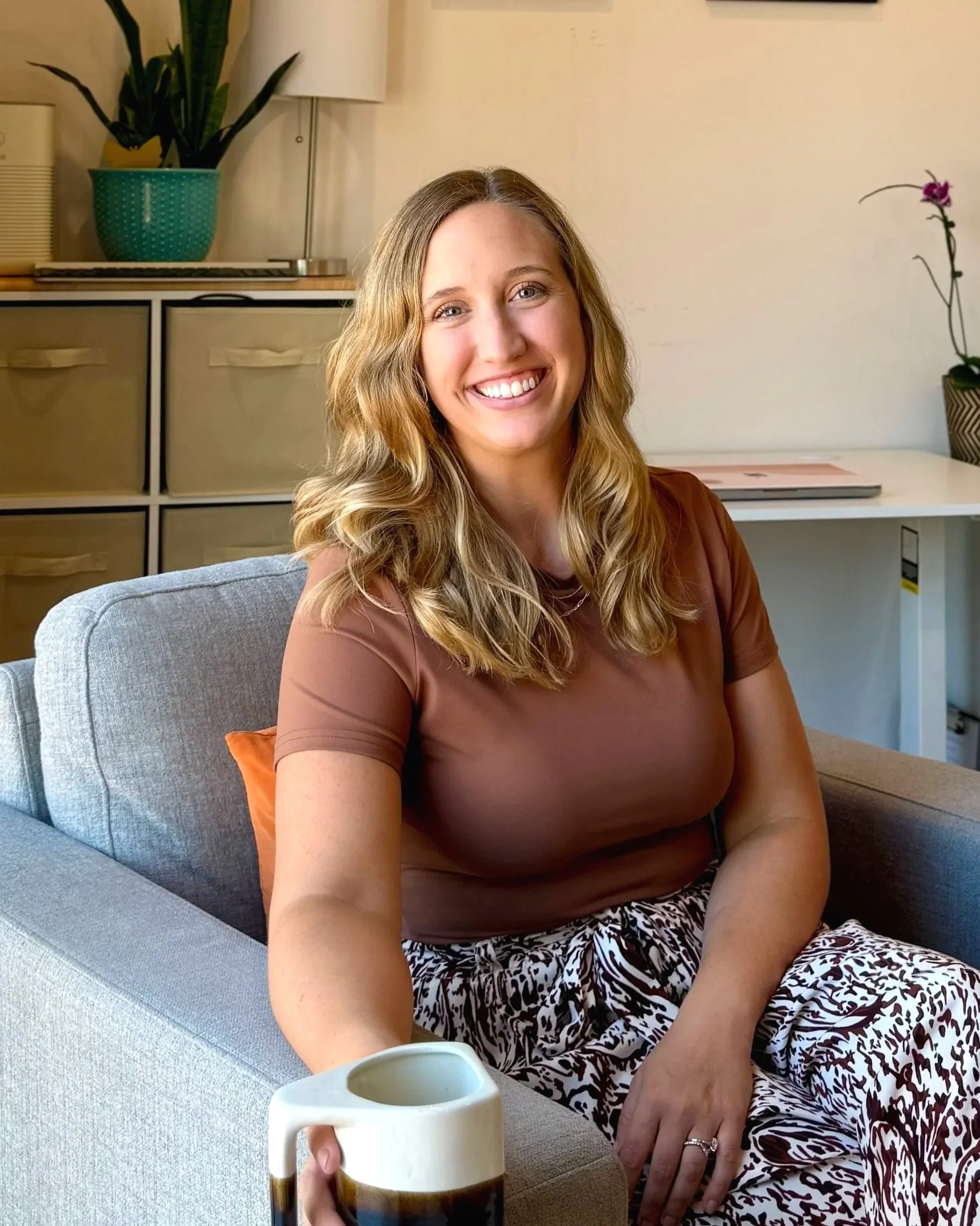 A woman sitting on a light gray sofa, smiling, holding a ceramic mug, with a plant, a lamp, a drawer, and a desk with a potted orchid in the background.