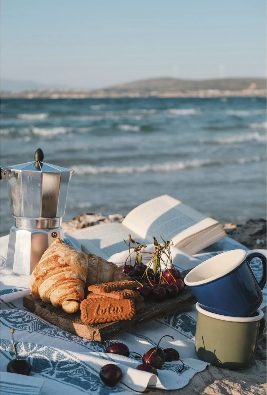 A seaside picnic setup with croissants, cherries, cookies, a French press, cups, and an open book on a cloth, with the ocean waves and distant shoreline in the background.