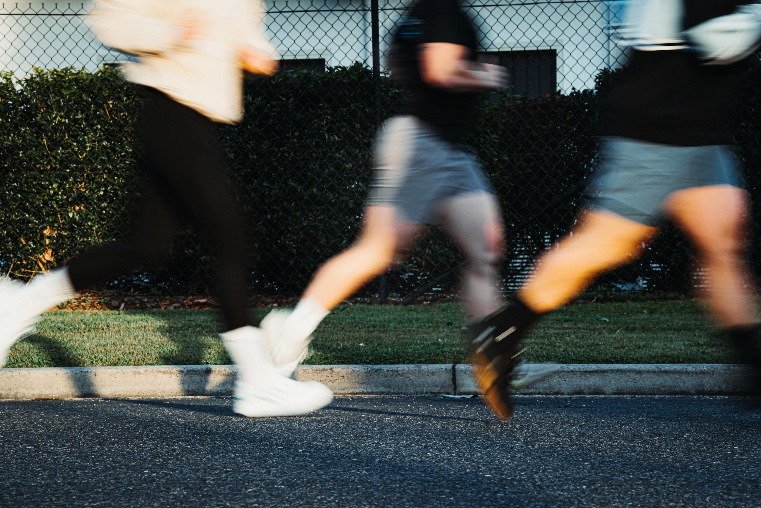 Blurred motion of three people running outdoors on pavement, with a black fence and bushes in the background.