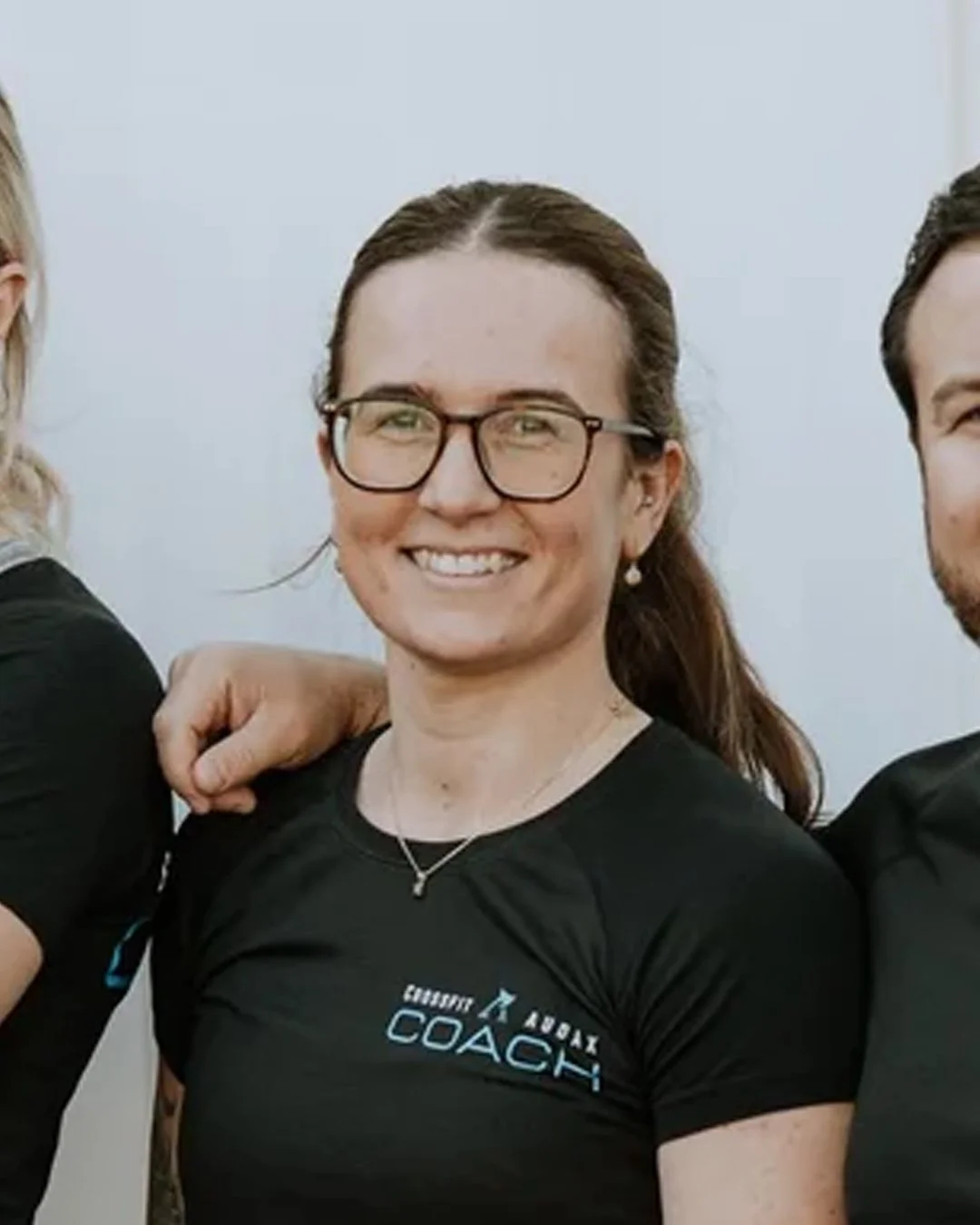 A woman with glasses and long brown hair smiling, wearing a black coaching T-shirt. She is surrounded by people with their arms around her shoulders, indicating camaraderie.
