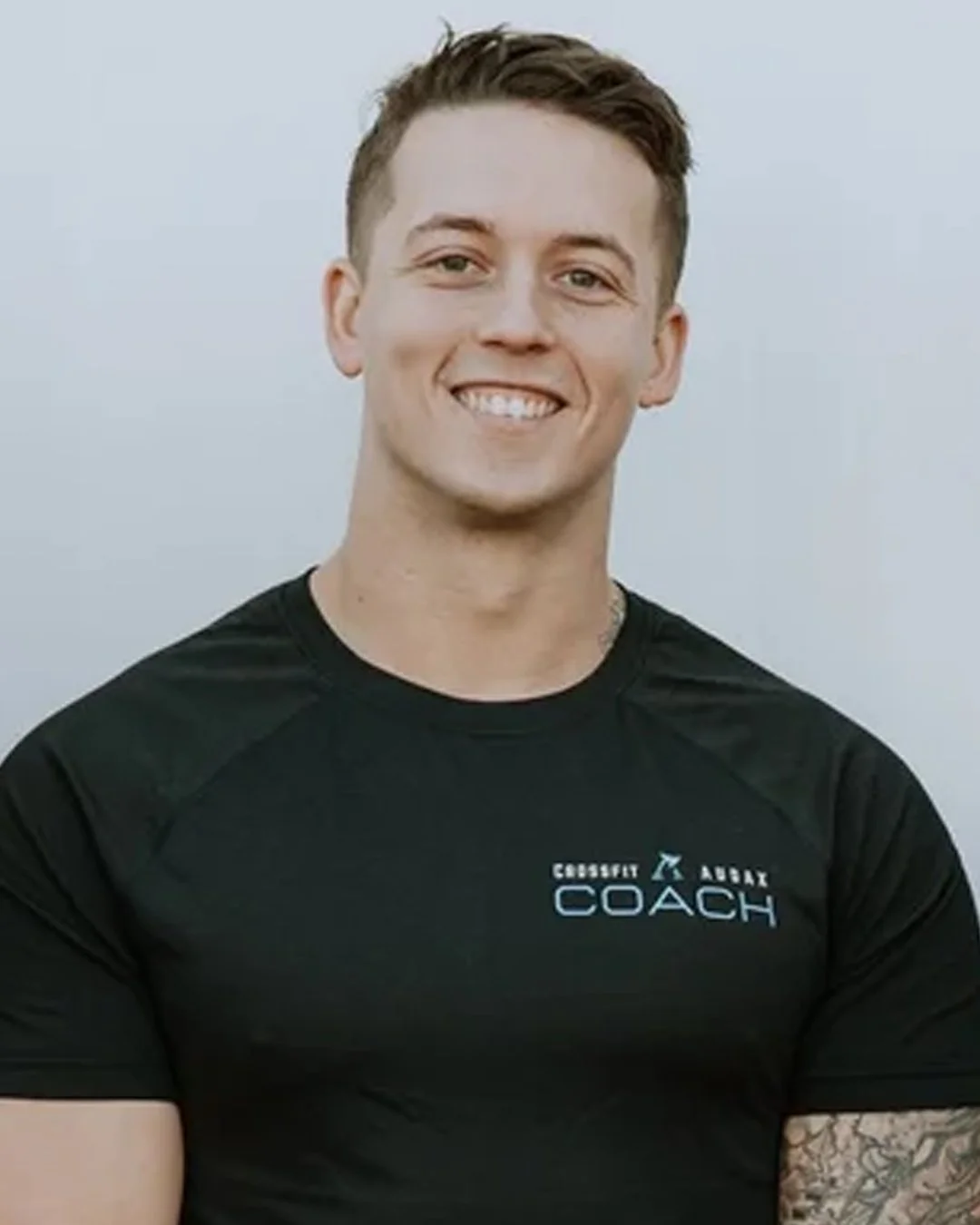A young man smiling while wearing a black athletic shirt with the words 'CrossFit Army Coach' on it.