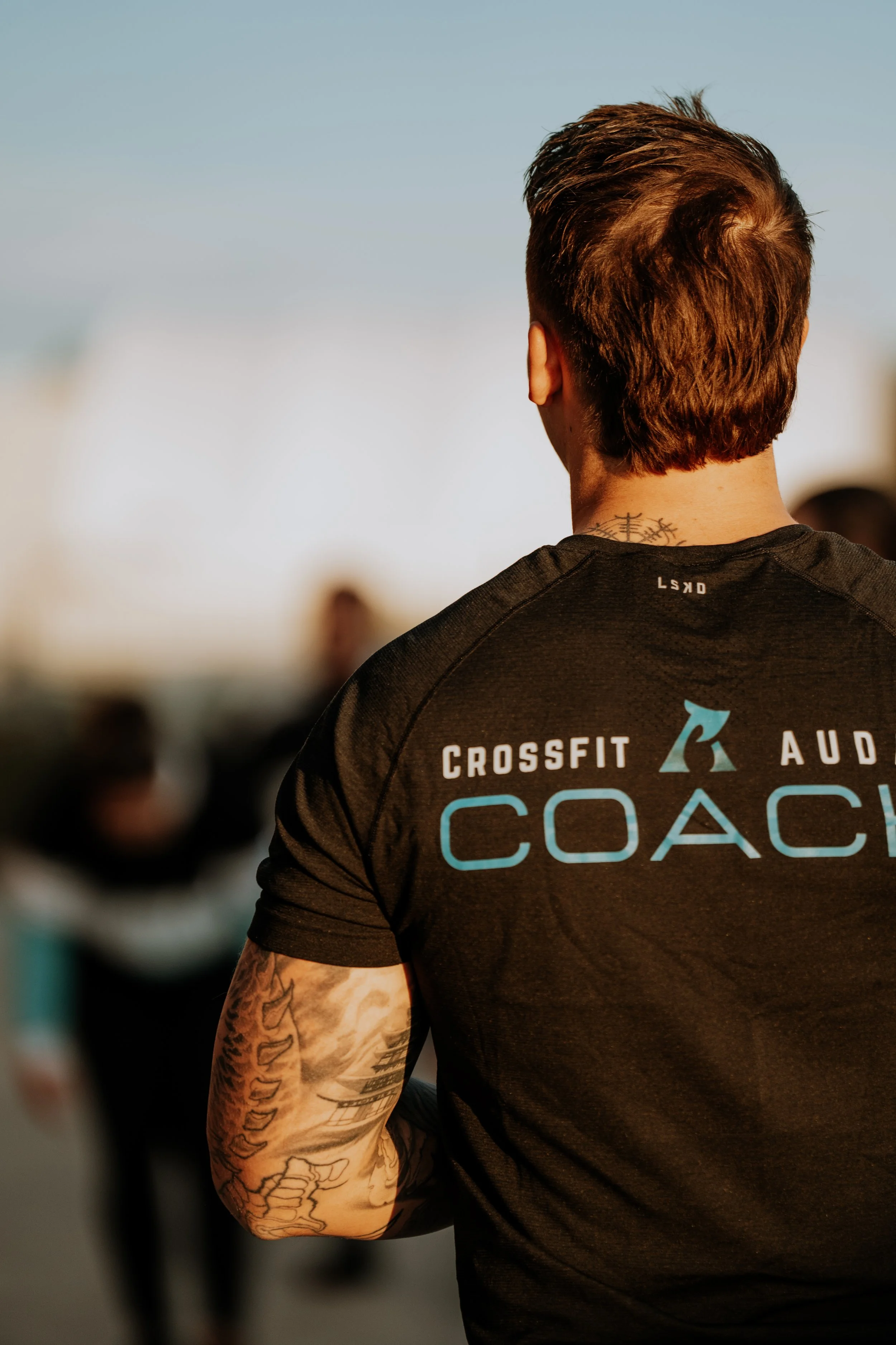 Back of a male CrossFit coach wearing a black shirt with blue and white lettering, standing outdoors with people blurred in the background.