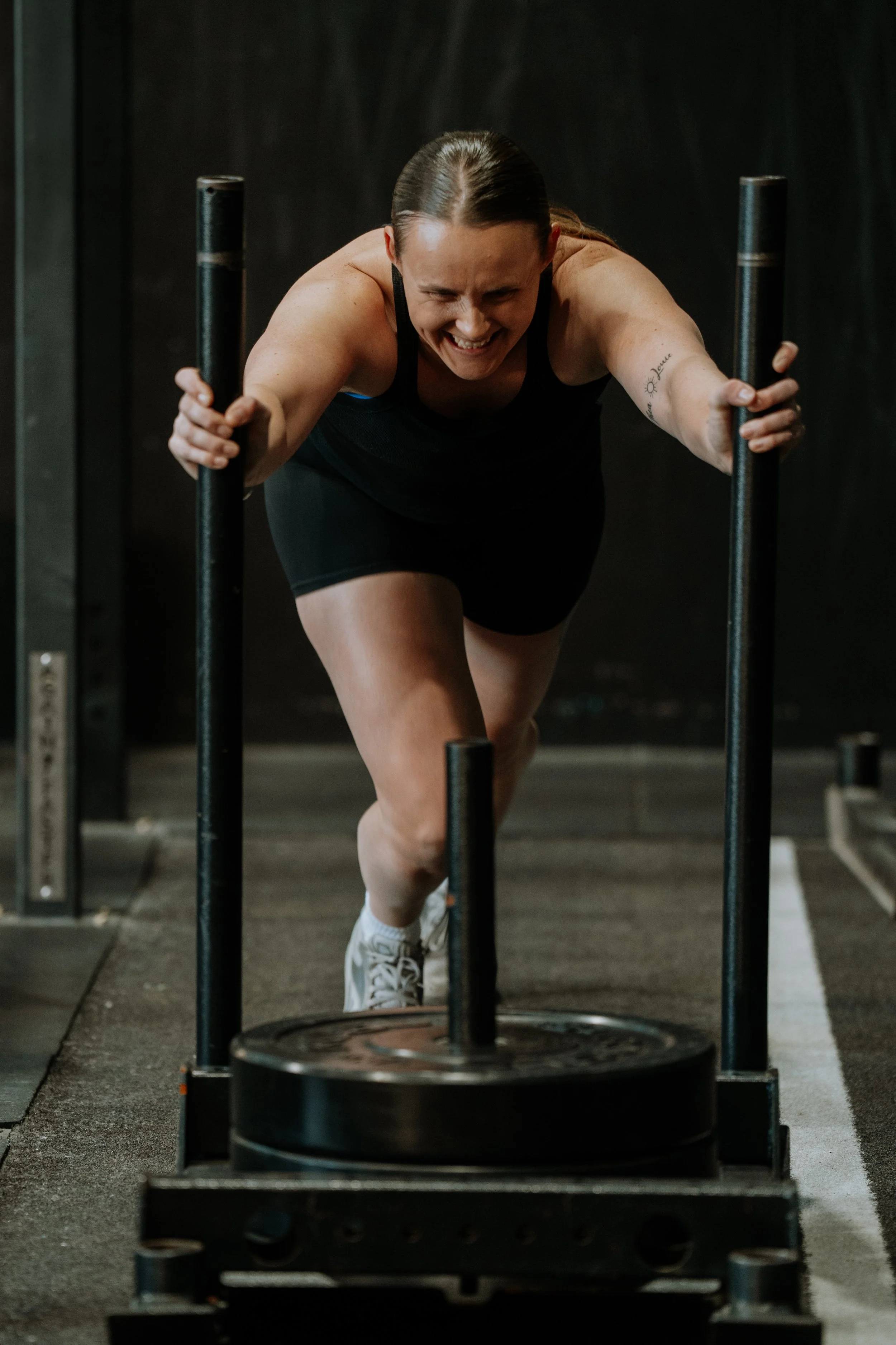 A woman wearing a black workout outfit and white sneakers pushing a weighted sled in a gym.
