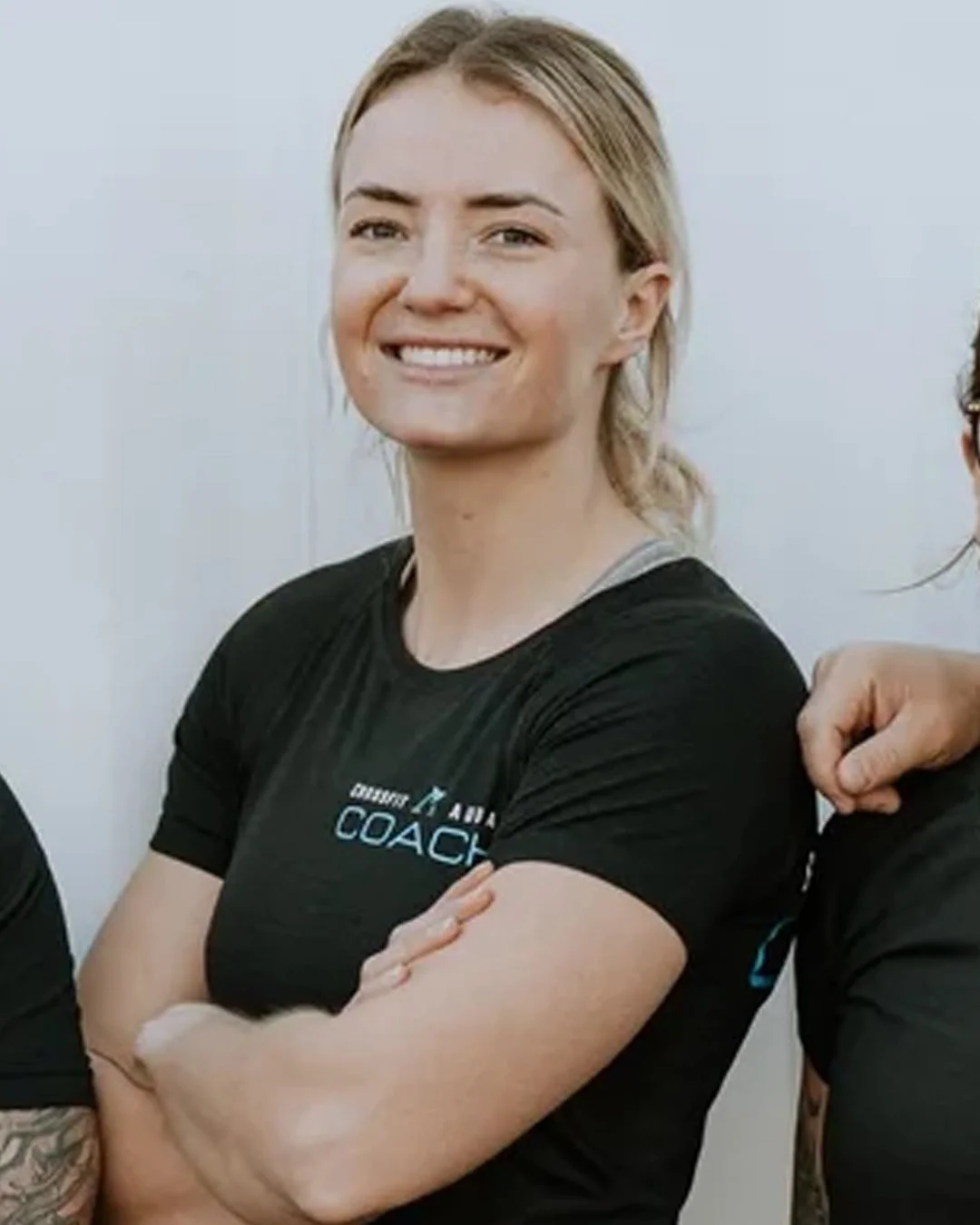 A woman with blonde hair smiling, crossed arms, wearing a black T-shirt with text that appears to say 'Coach' and some other smaller text, against a plain light-colored background.
