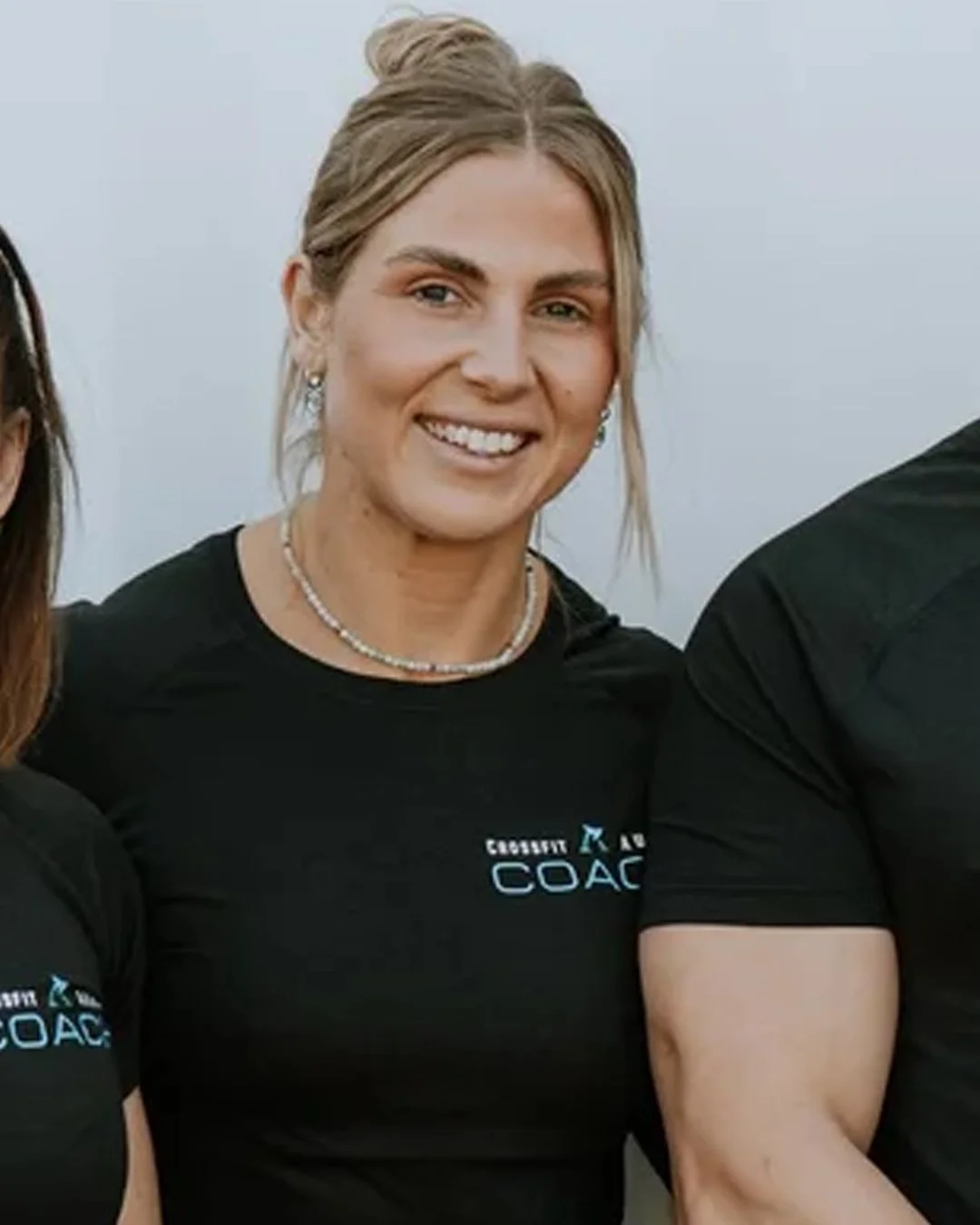 A smiling woman with blonde hair styled in an updo, wearing a black shirt with a logo that says 'COACH', a pearl necklace, and earrings, standing outdoors with a clear sky in the background.