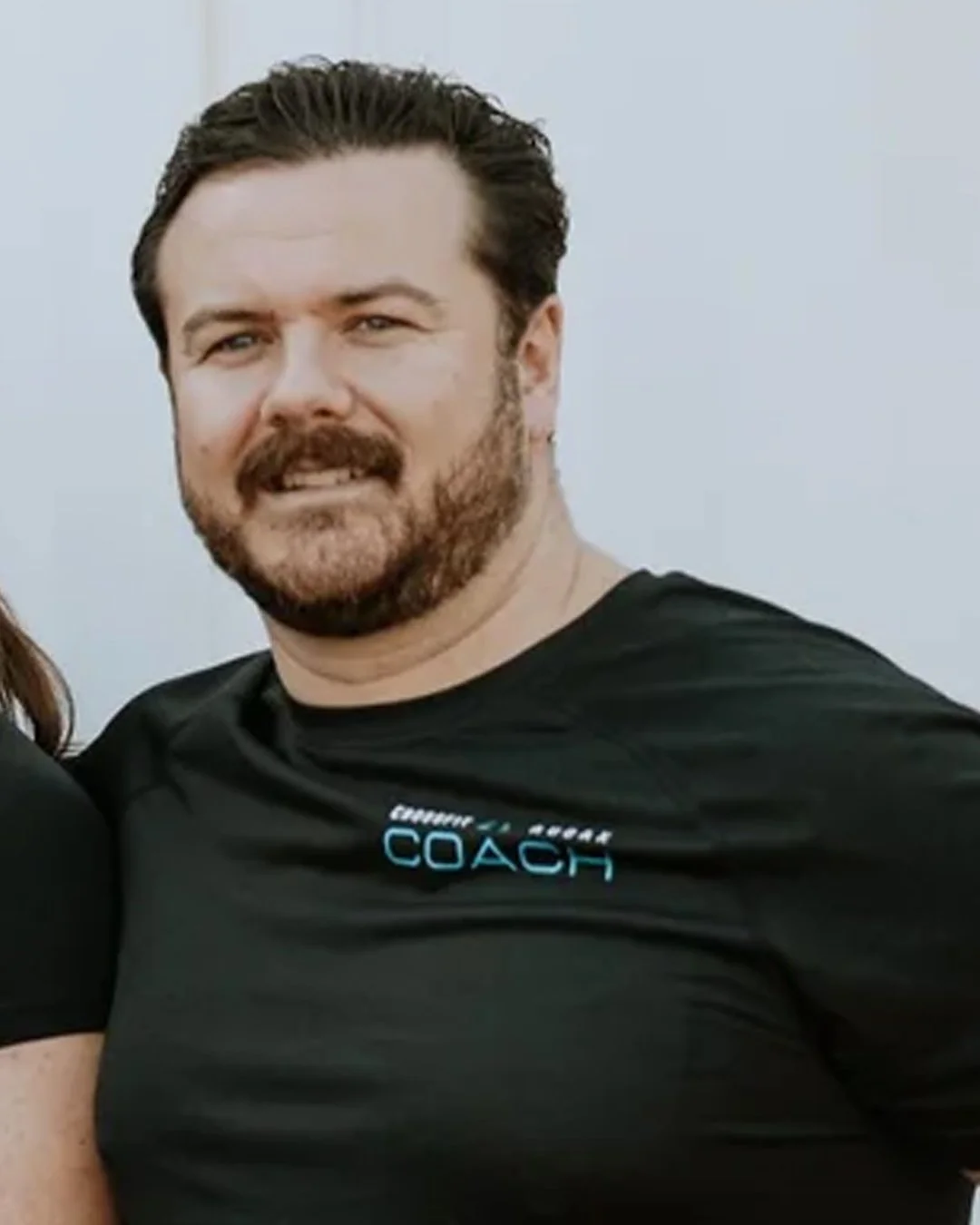 Man with dark hair, beard, and mustache wearing a black athletic shirt with the word 'COACH' on it.