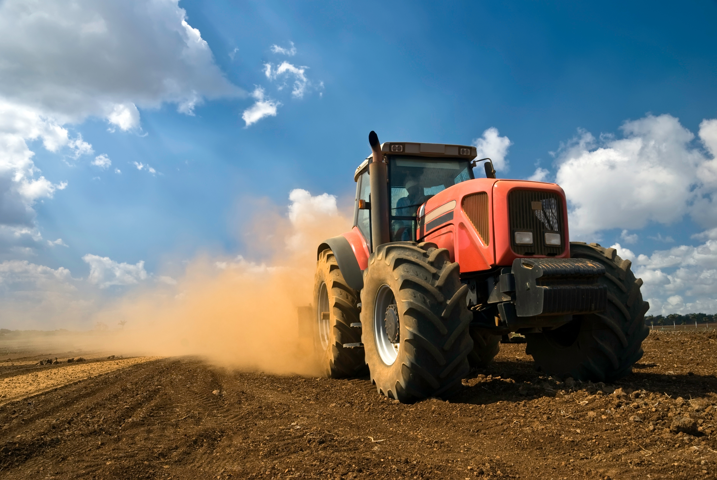 A red tractor working on a farm field, kicking up dust under a blue sky with scattered clouds.