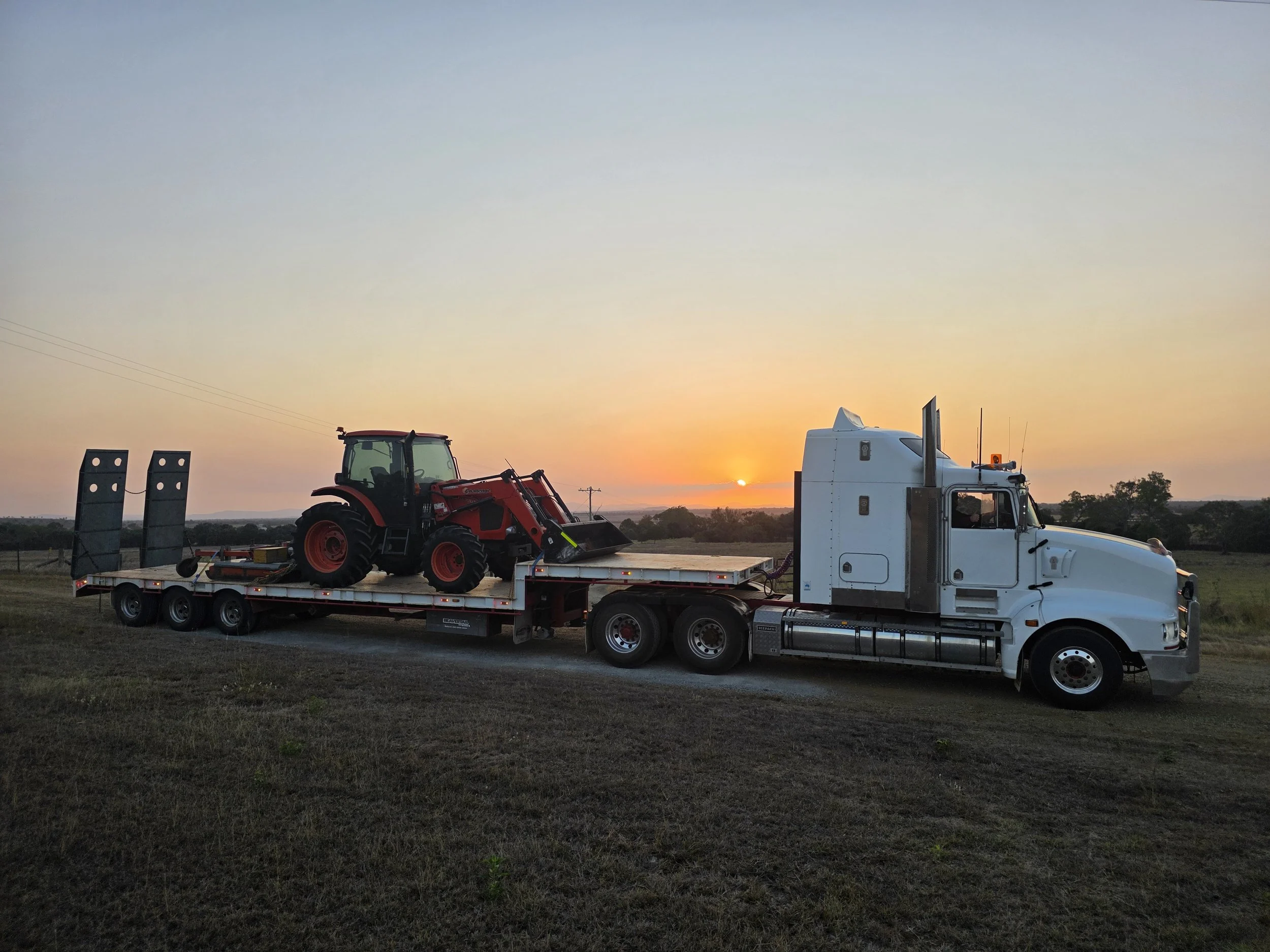 A flatbed truck transporting a red tractor at sunset, with open fields and trees in the background.
