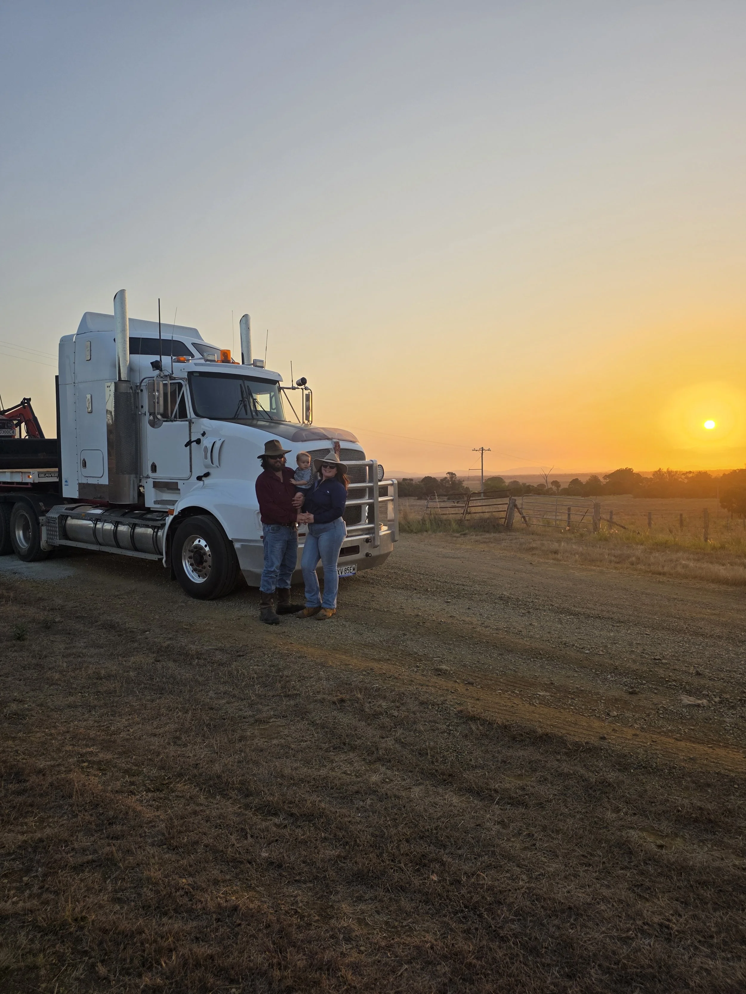 A family of three standing in front of a large white semi-truck on a dirt road during sunset, with open fields and a fence in the background.