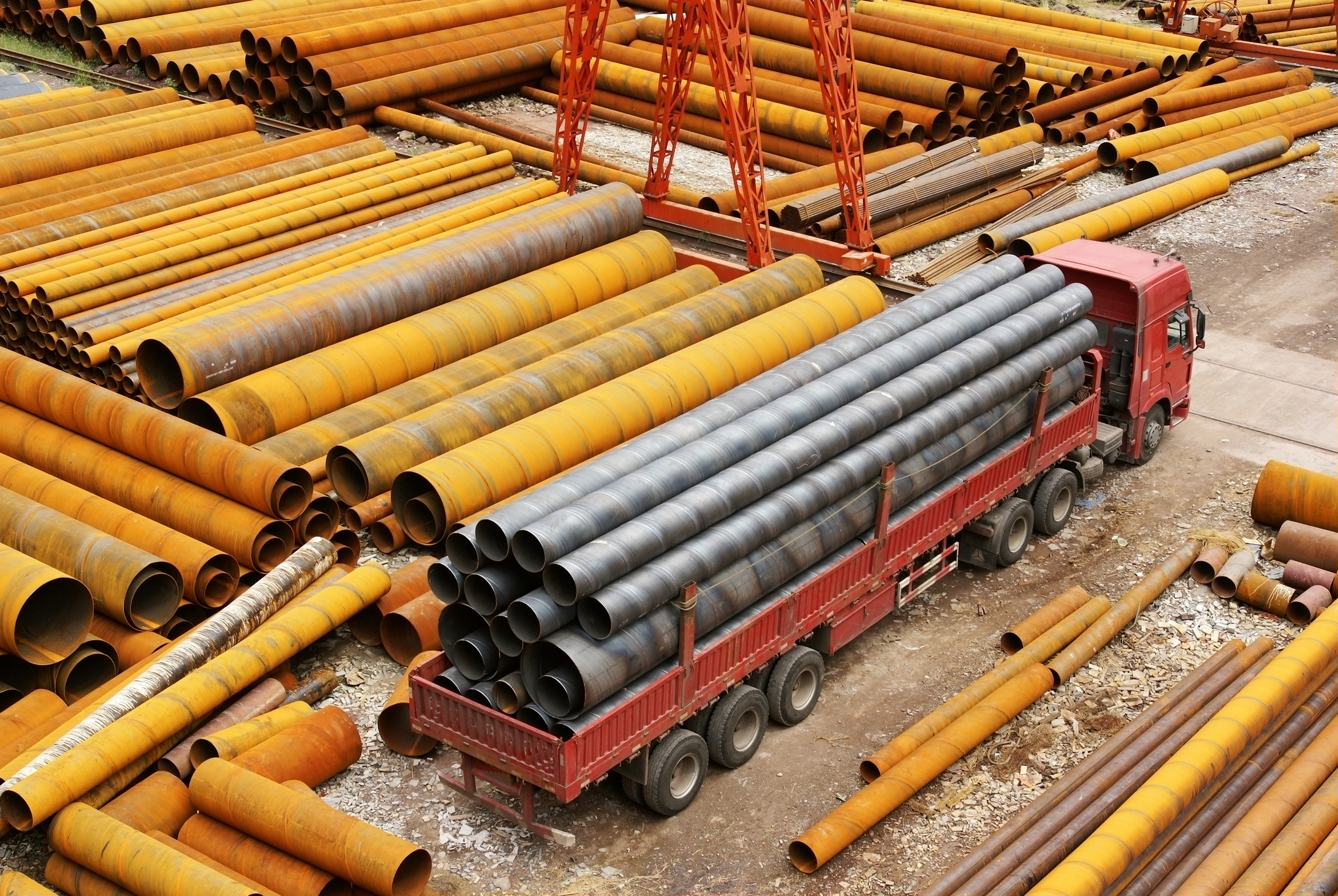 Red truck carrying large gray metal pipes parked among stacks of yellow and rust-colored metal pipes in an industrial yard.