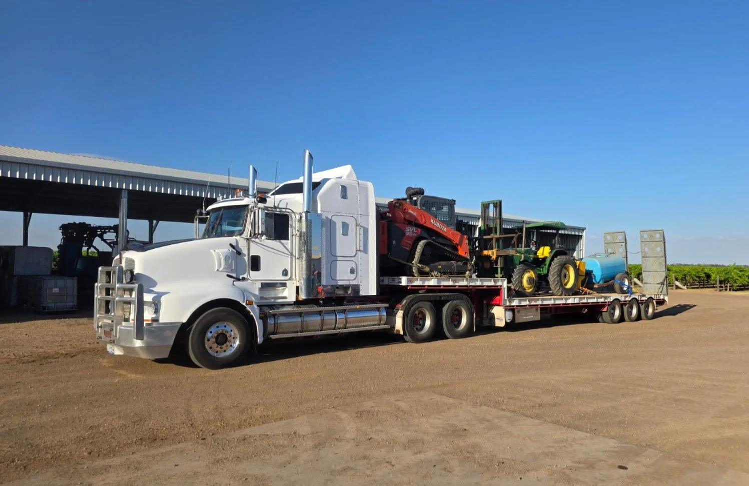 White flatbed truck carrying a red compact excavator, a green tractor, and a blue water tank parked on a dirt lot under a blue sky.