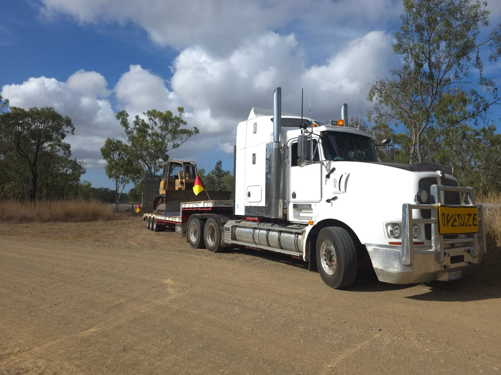 A white semi-truck with a flatbed trailer carrying a yellow construction vehicle on a dirt road, with trees and a partly cloudy sky in the background.