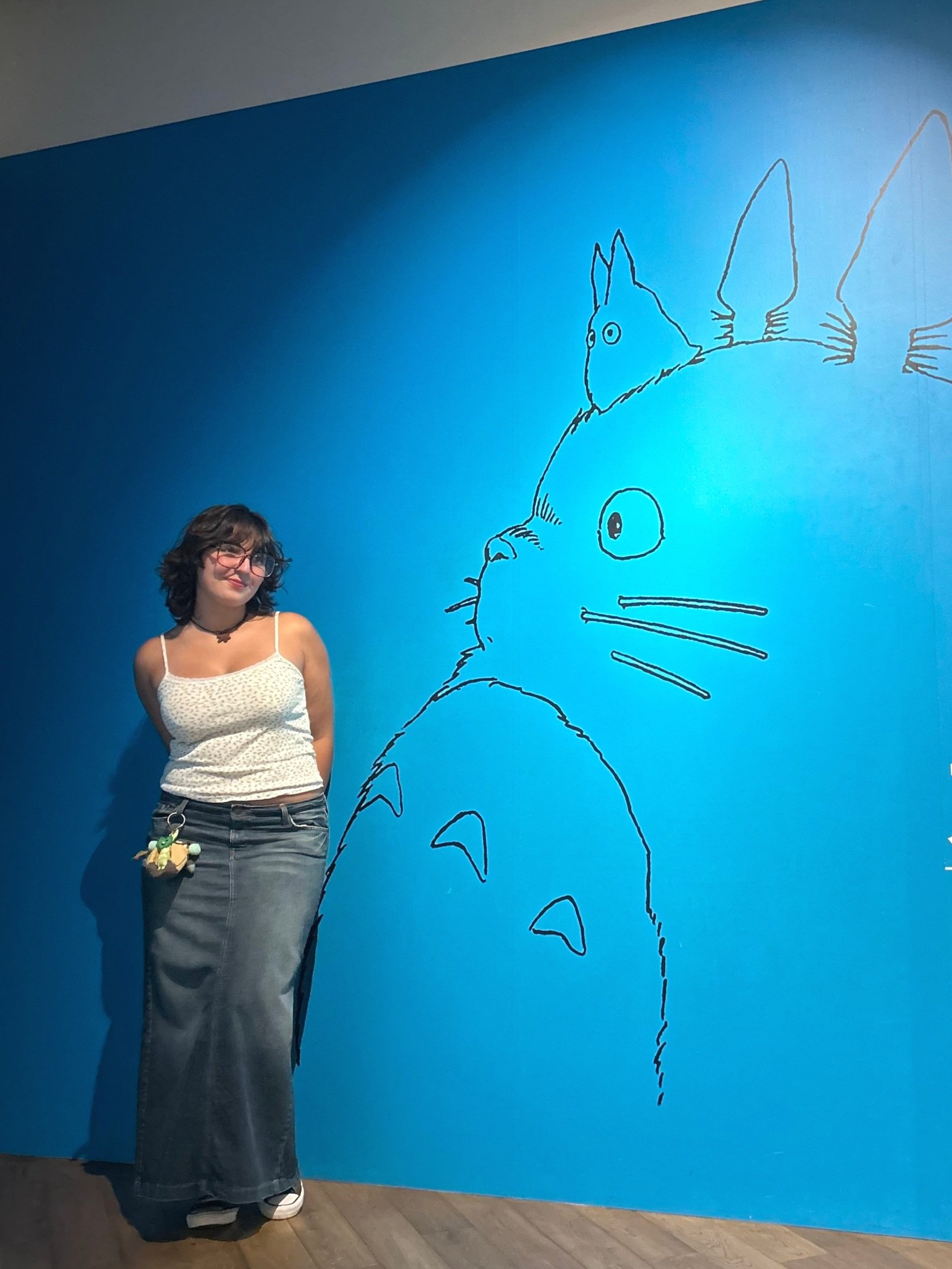 A young woman with glasses, a white tank top, and gray skirts stands against a blue wall with a large, simple line drawing of Totoro, a character from Japanese animation, and a smaller creature on his head.