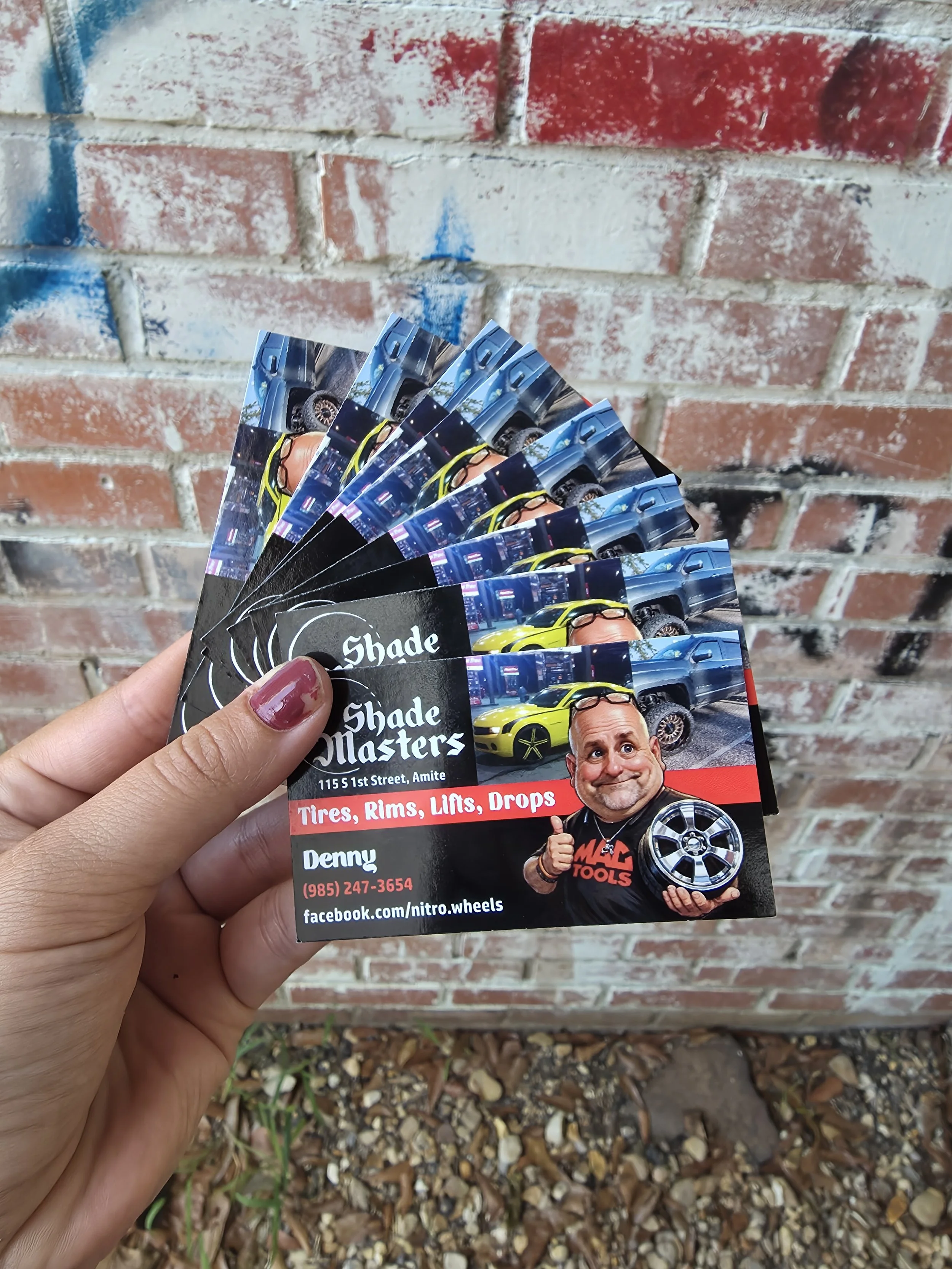 Hand holding a stack of business cards with info about car wheels and customization, in front of a red and white brick wall.