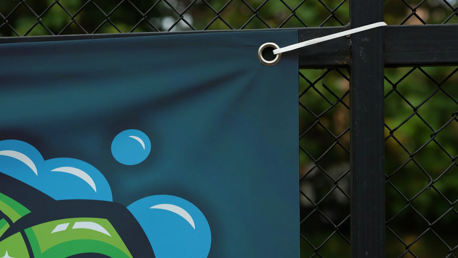 Close-up of a black metal fence with a sign attached by a white zip tie. The sign features a partial graphic of clouds and green landscape.