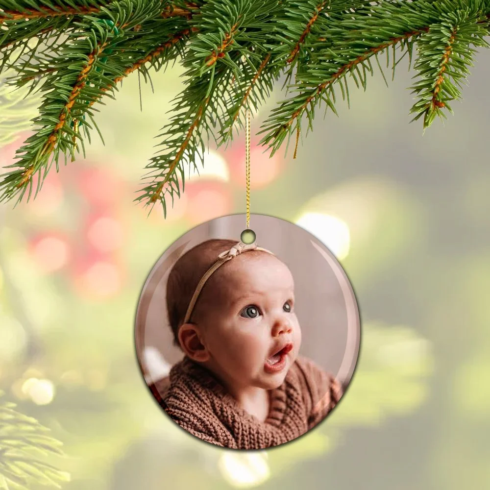 A Christmas tree branch with a hanging ornament featuring a close-up photo of a surprised baby with big eyes, wearing a brown sweater and a headband.