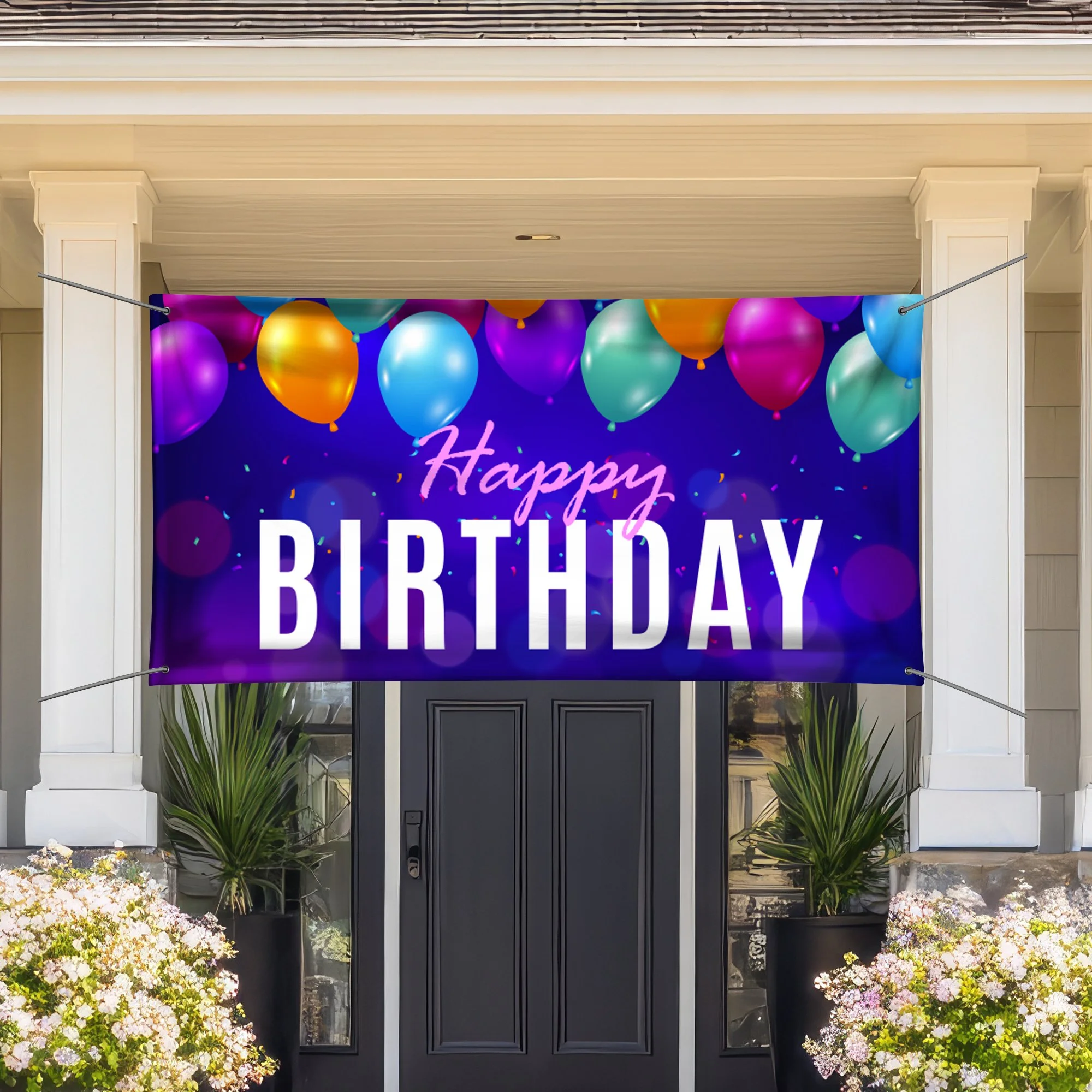 A house with a porch decorated for a birthday celebration, featuring a large sign with balloons that reads 'Happy Birthday.' The house has white trim, black door, and garden plants with blooming flowers.