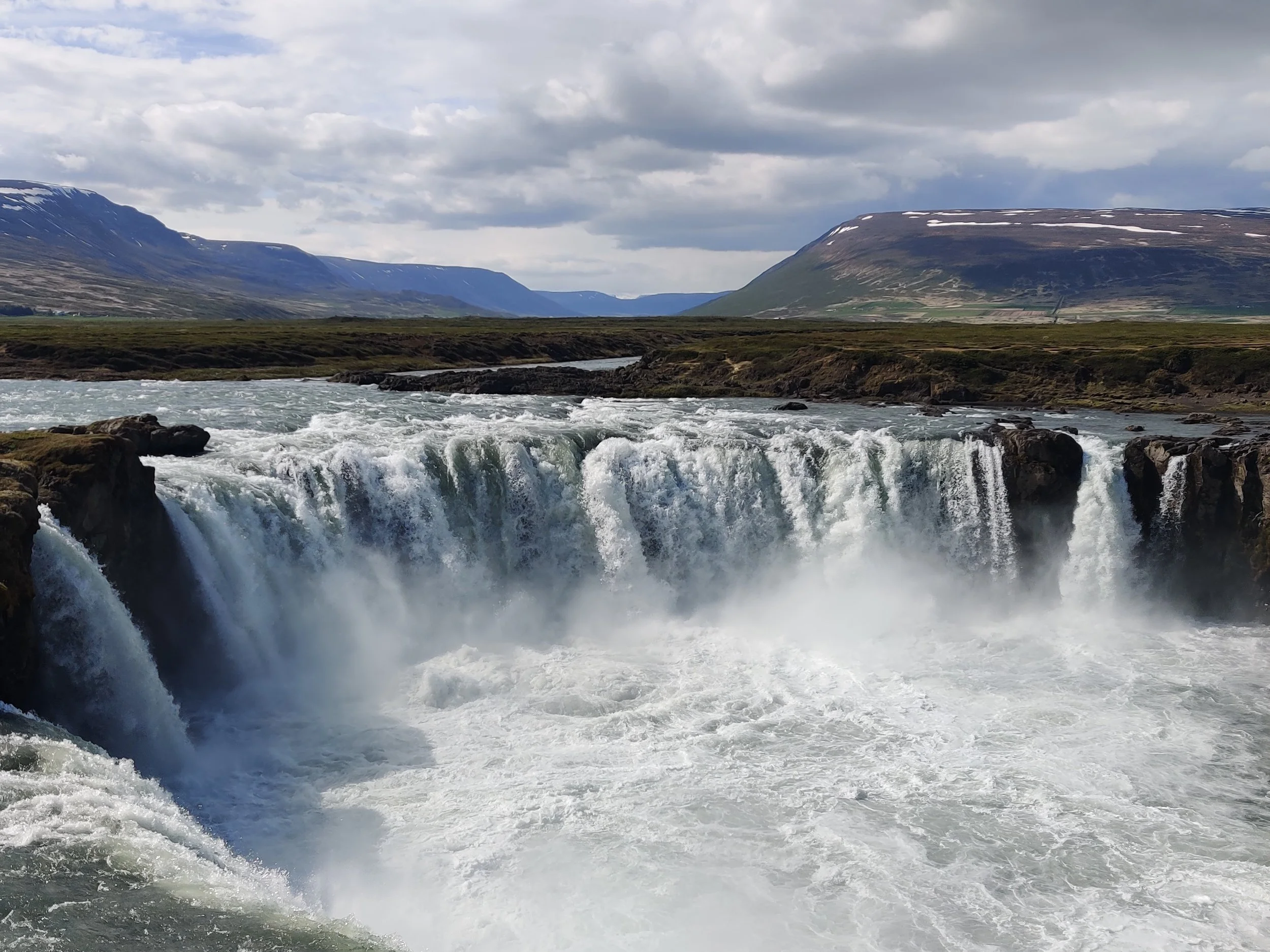 Goðafoss Waterfall in Iceland - powerful waterfall flowing into a river surrounded by mountains under a cloudy sky.