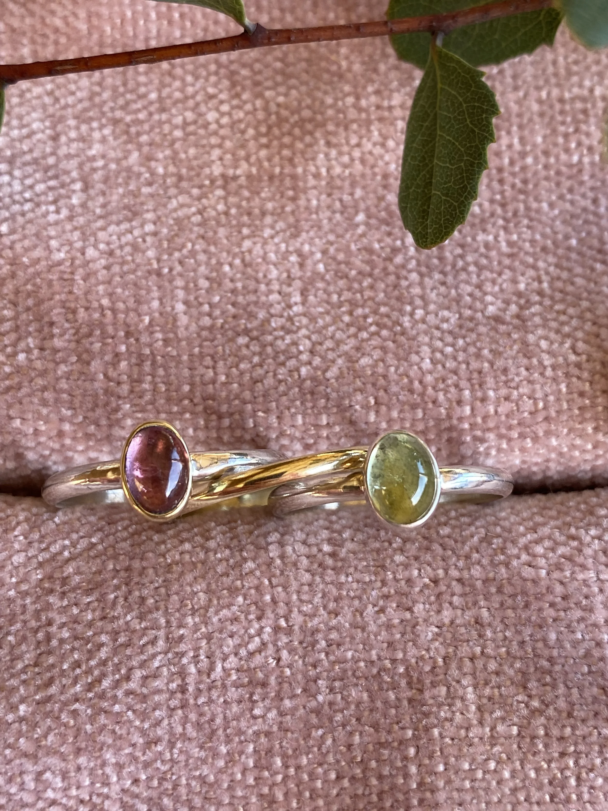 Two intertwined rings with oval gemstones, one with a red gemstone and the other with a green gemstone, resting on a pink textured fabric with a green leaf above.