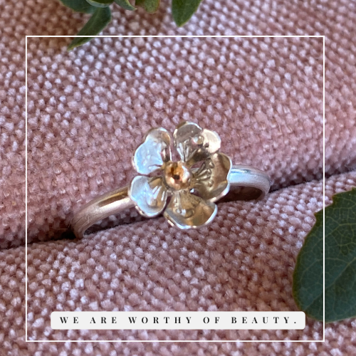 Silver ring with a flower design featuring gold and silver petals, resting on pink fabric with green leaves nearby.