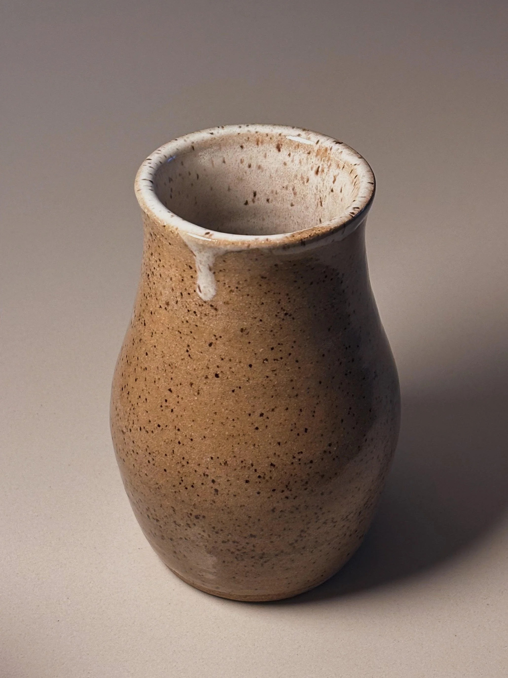 A small, beige, speckled ceramic vase with a narrow opening and a slightly flared rim, sitting on a neutral surface.