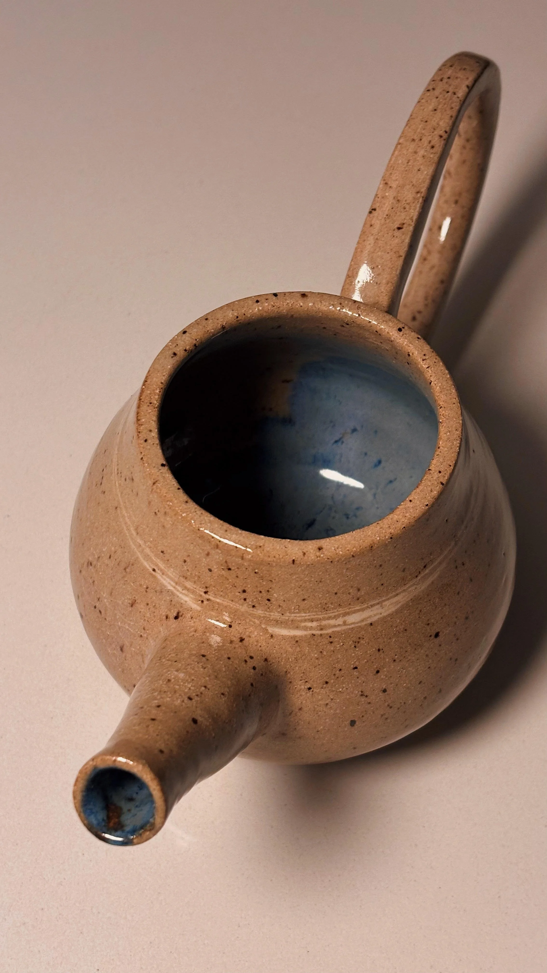 A tan, speckled ceramic teapot with a handle and spout, placed on a light surface.
