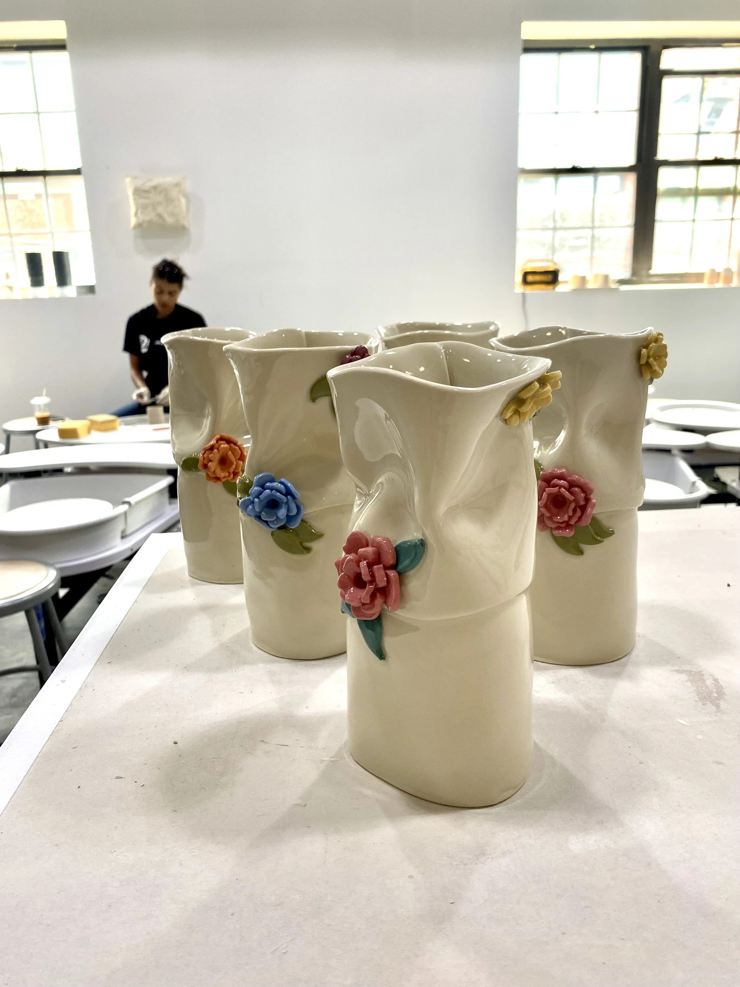 Ceramic vases with floral designs displayed on a white table in a bright room, with a person working in the background.