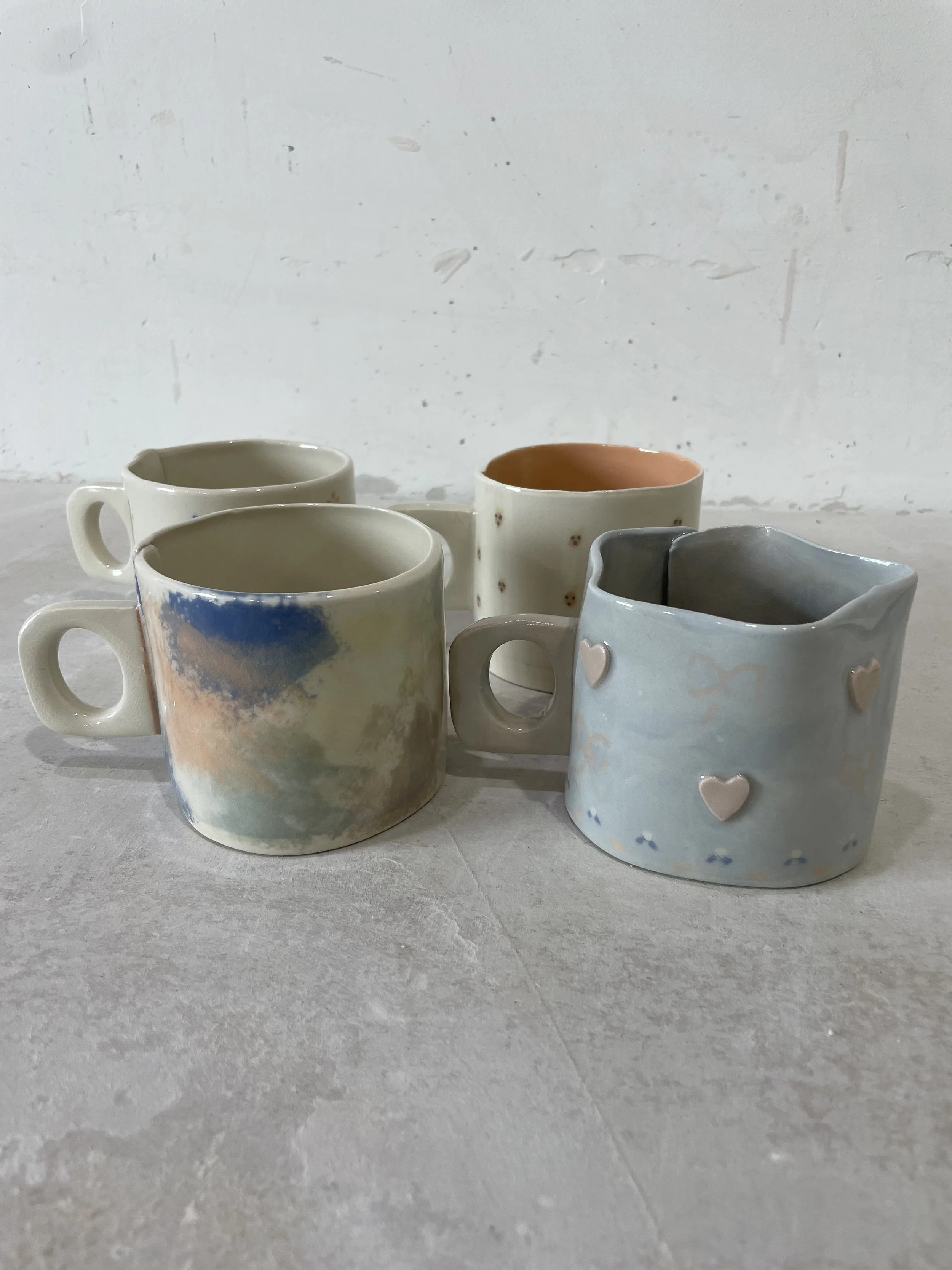Five handmade ceramic mugs with various painted designs arranged on a light-colored surface against a plain wall.