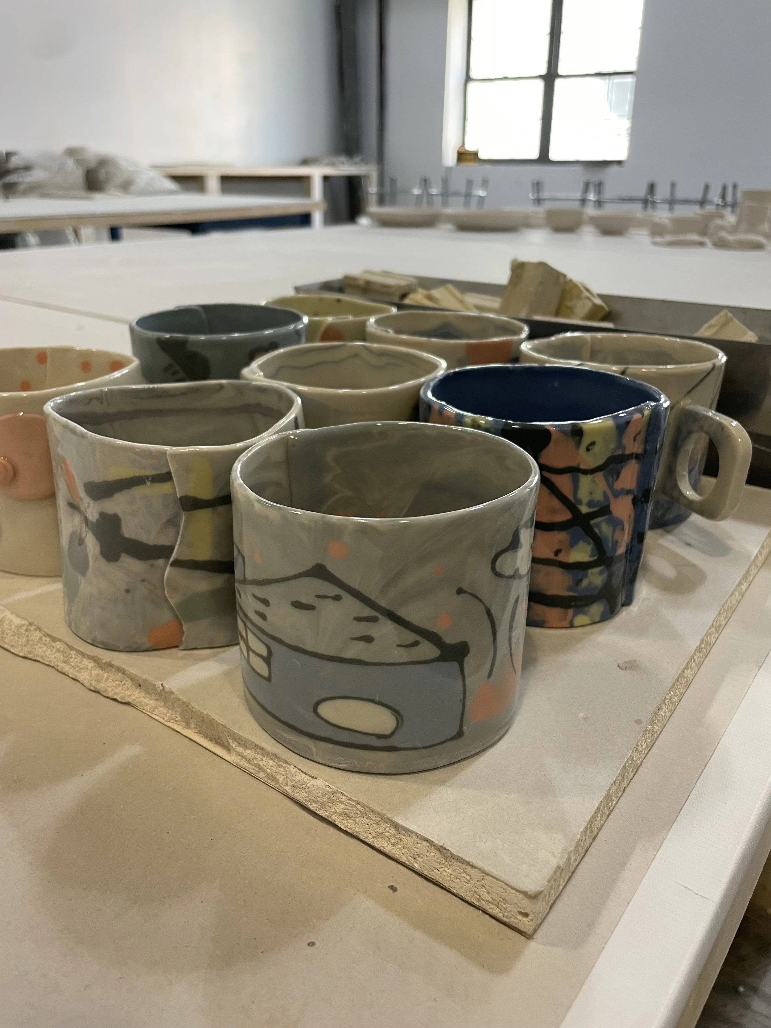Handcrafted ceramic mugs with abstract designs arranged on a work table in a pottery studio.