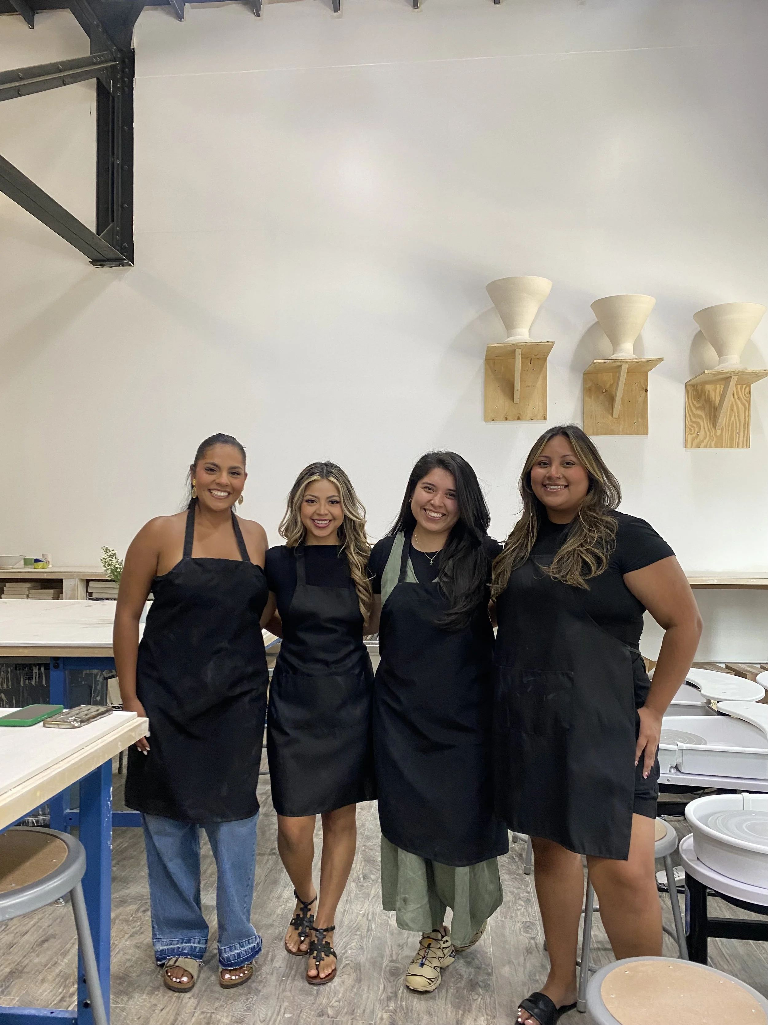 Four women in black aprons standing together in a pottery studio, smiling for the photo.