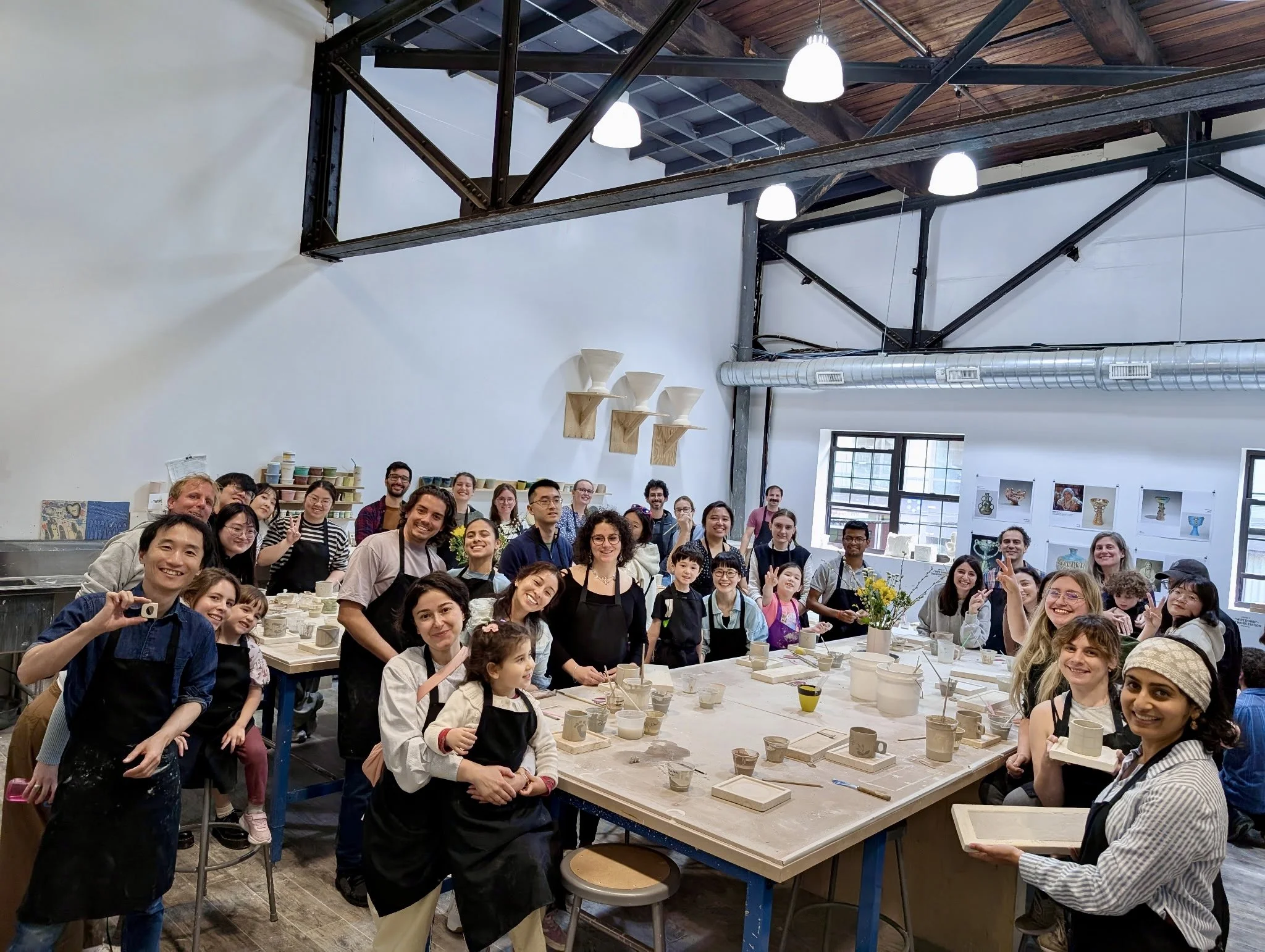 Group of people in a pottery studio, some holding pottery items, surrounded by pottery supplies and artworks, smiling for the camera during a class or workshop.