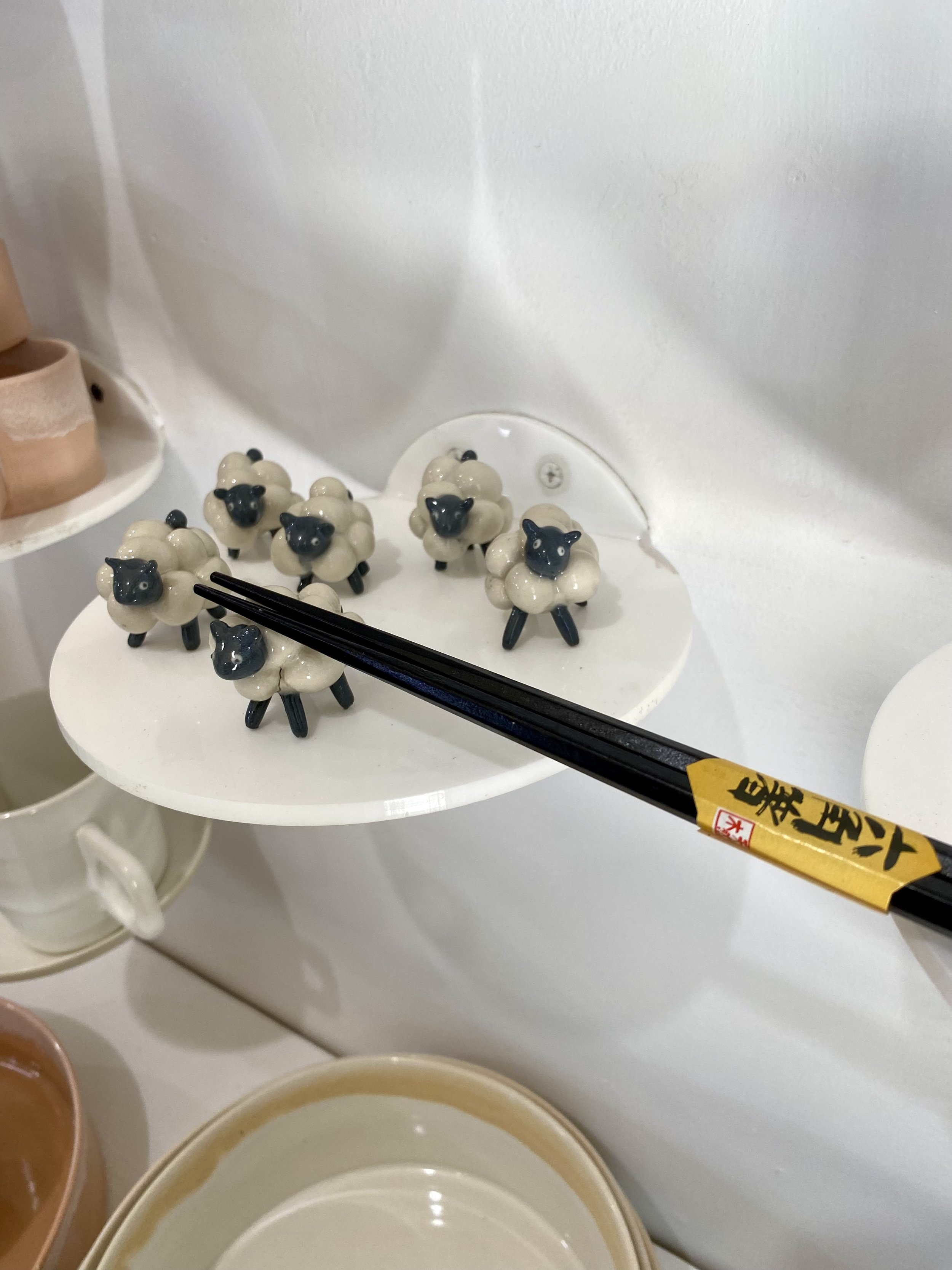 Small ceramic figurines of black-faced sheep with white woolly bodies arranged on a white round display table, with black chopsticks resting on them. The background features a white wall and other ceramic items.