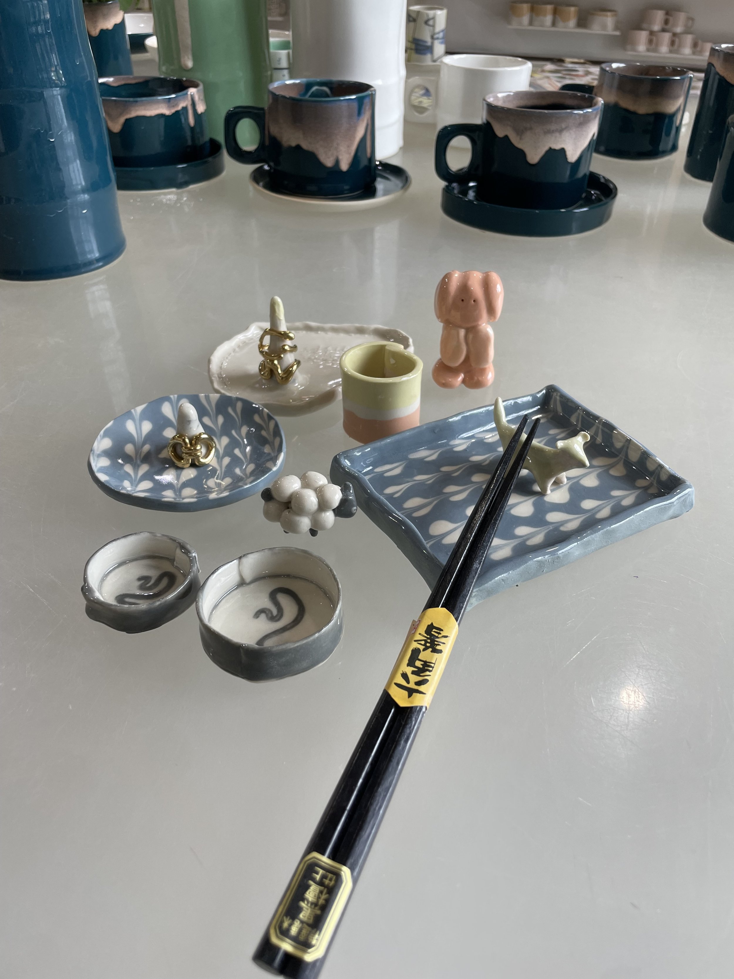 Decorative miniature ceramics and figurines on a white table, including a pink dog figure, small bowls, a striped cup, and chopsticks.