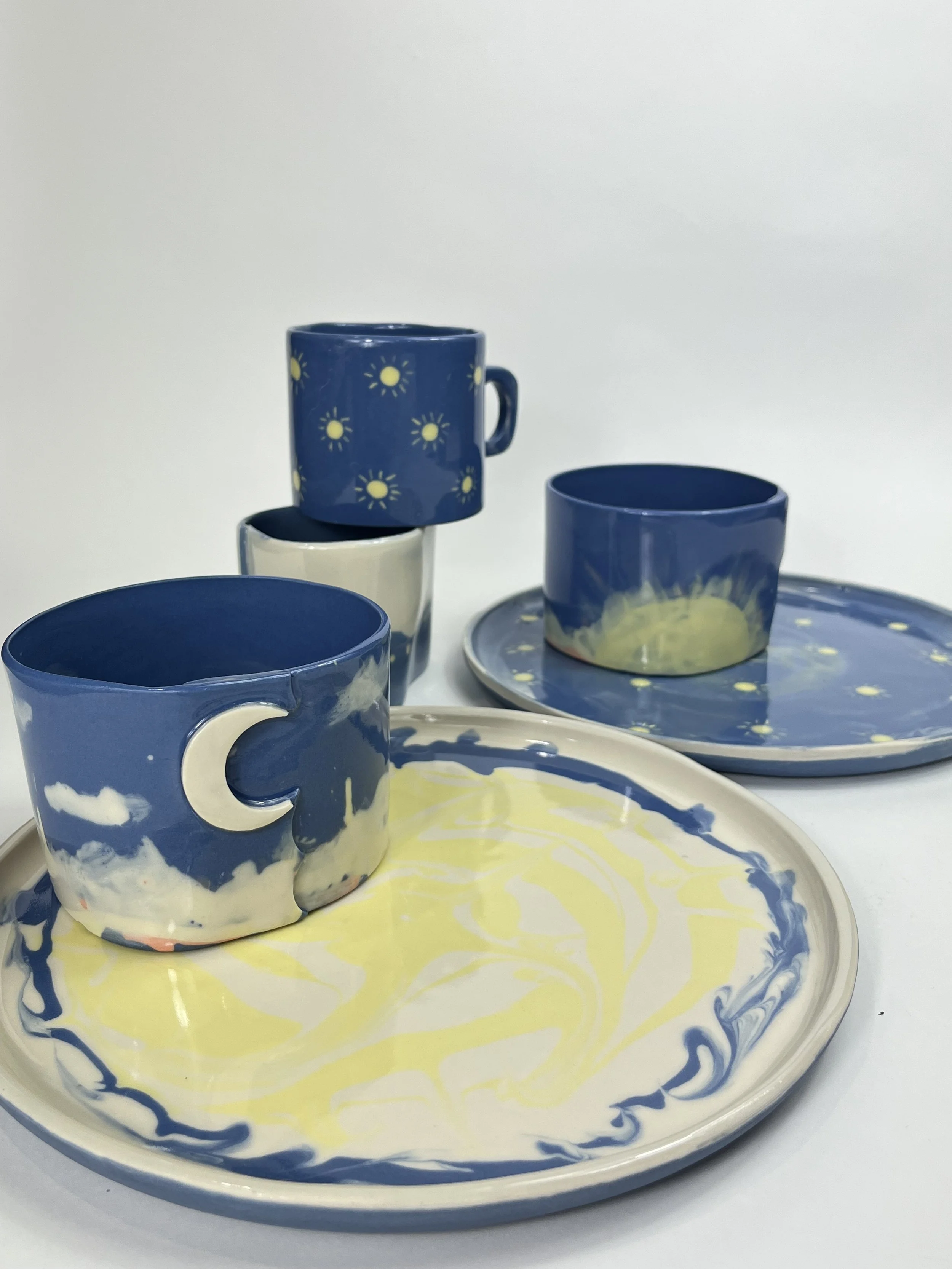 Set of ceramic tableware with a yellow and blue swirling pattern, including plates and cups with star and moon designs and a yellow sun motif.