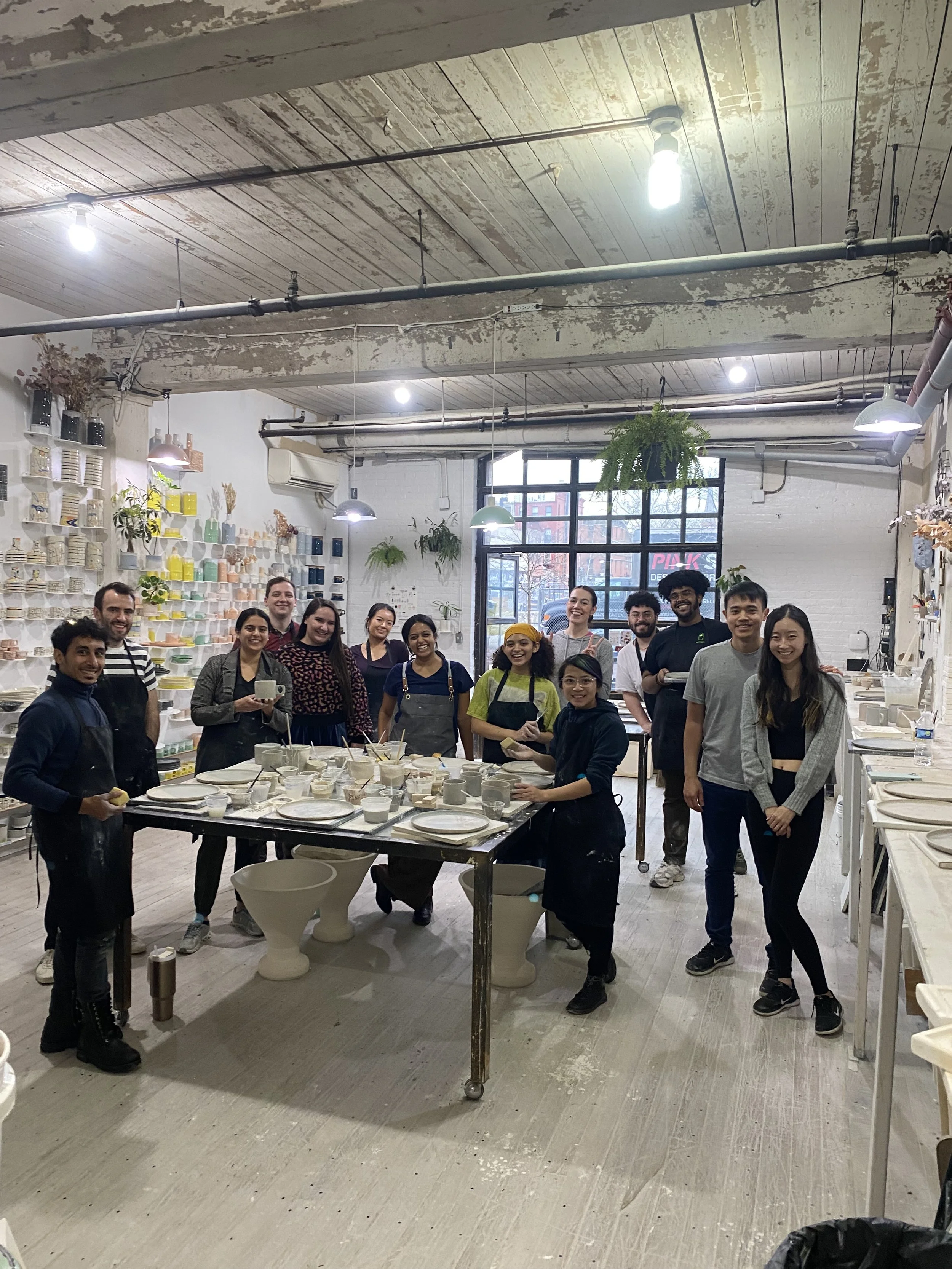 Group of people smiling and posing in a ceramics studio, surrounded by pottery and art supplies.
