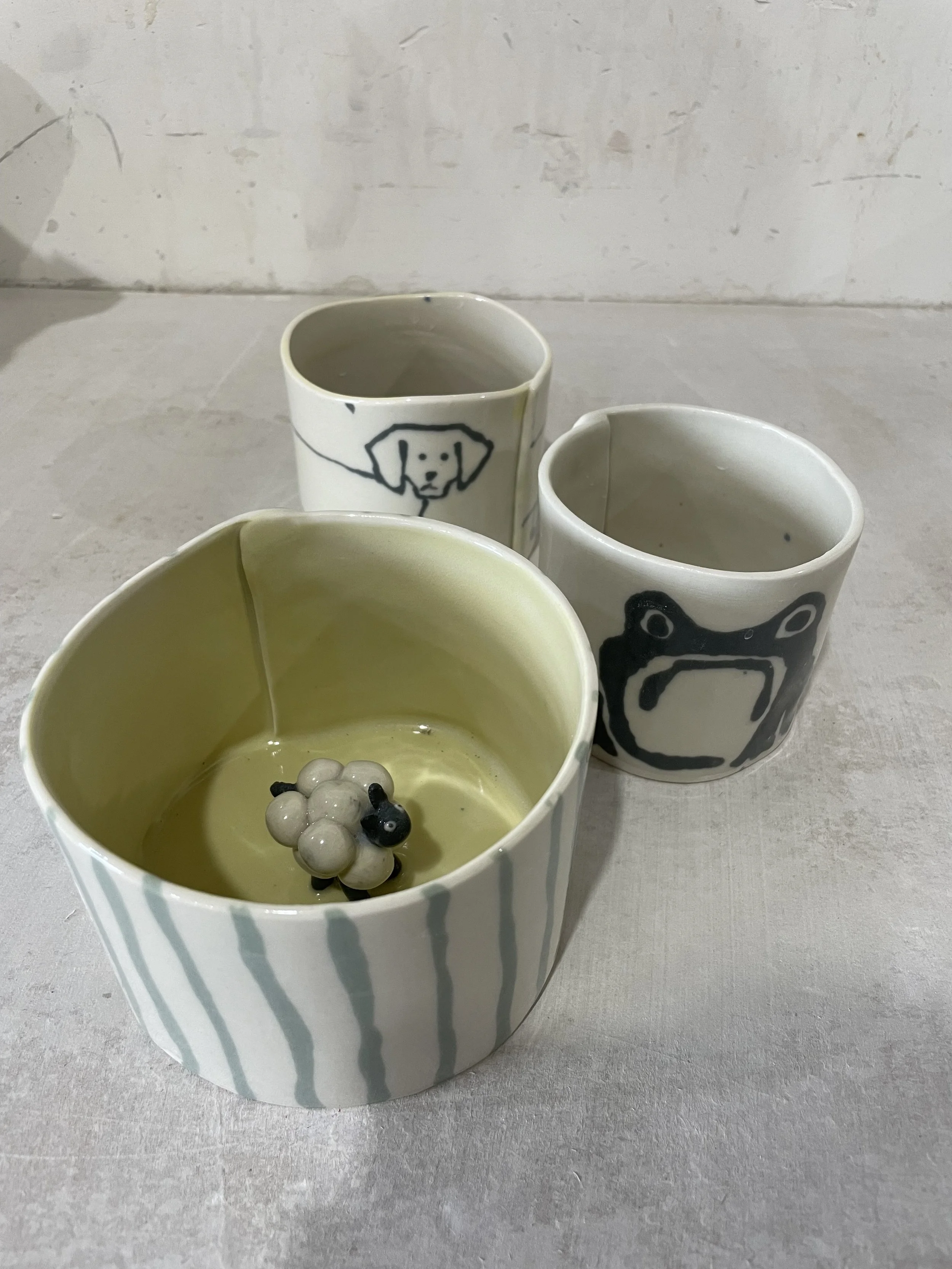 Three handmade ceramic cups with animal illustrations, one with a sheep figurine inside.