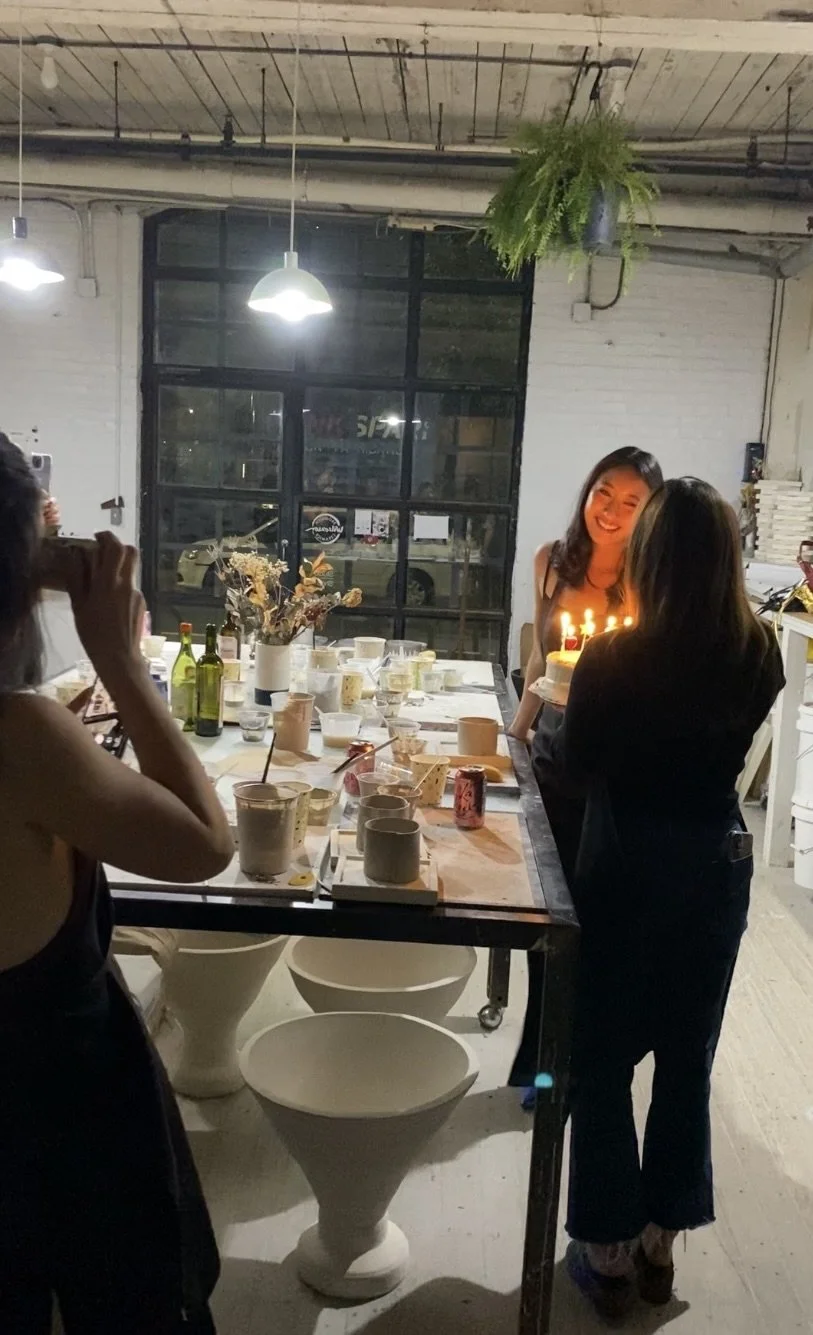 A group of women celebrating a birthday with a cake and candles in a modern industrial-style room, with a decorated table and ambient lighting.