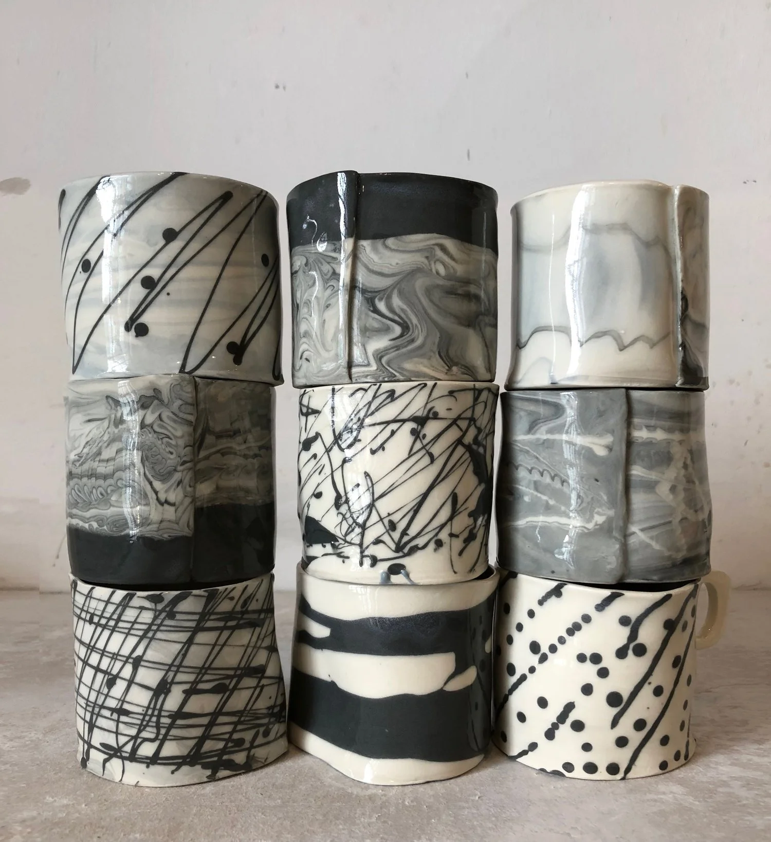 A set of nine black and white marbled and splattered ceramic mugs arranged in three rows on a concrete surface.
