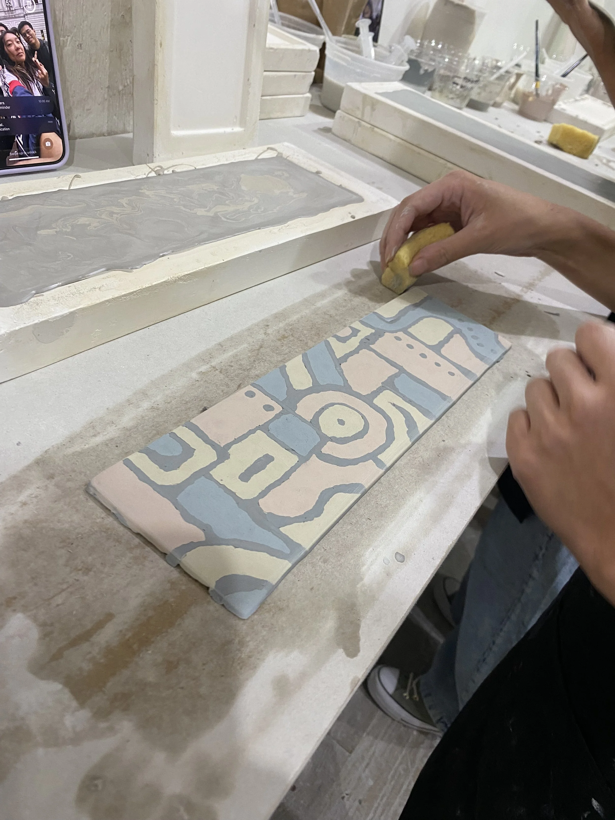 Person is sanding a colorful, patterned ceramic tile at a worktable in an art studio, with art supplies and trays of materials in the background.